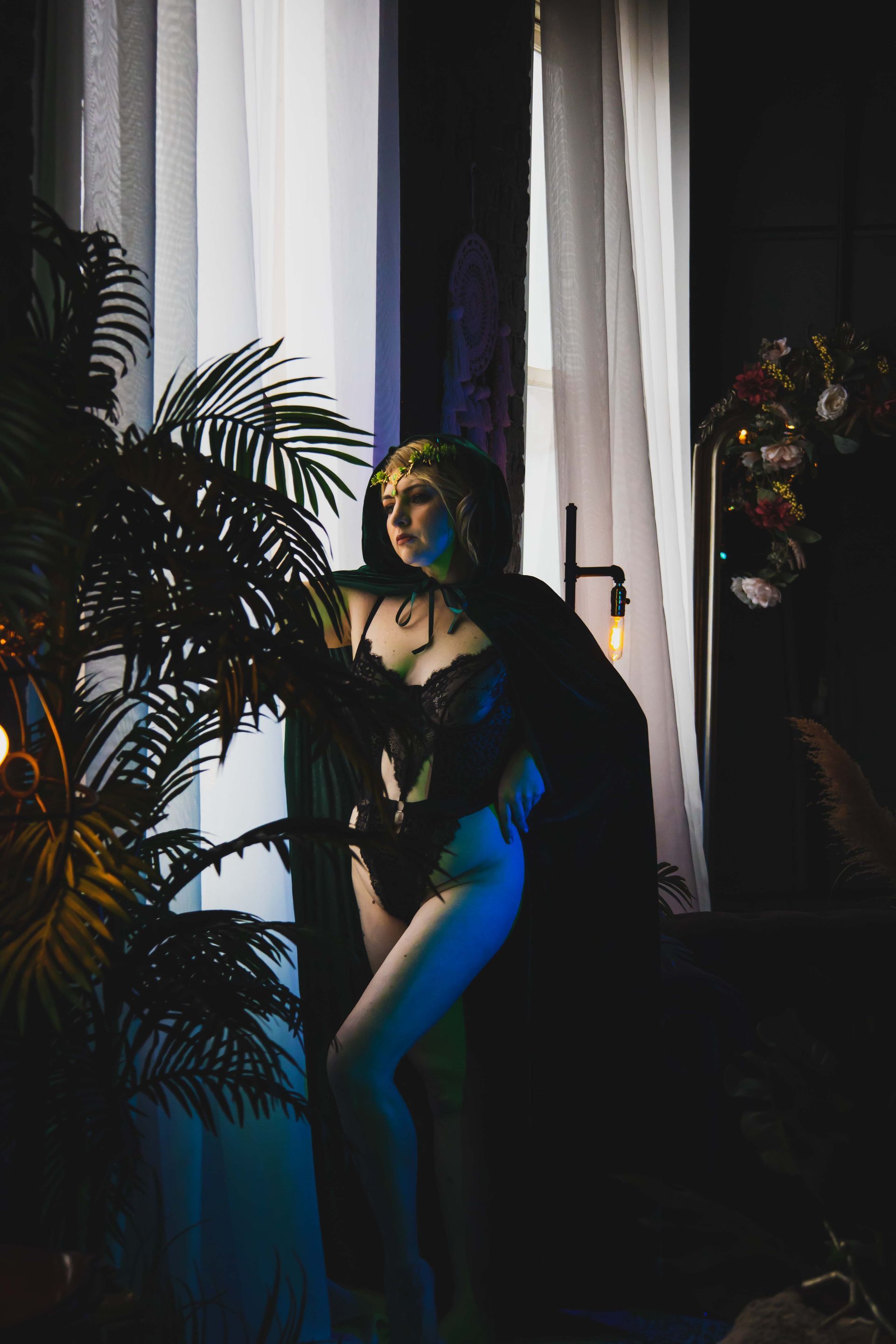 Woman in black lingerie and cloak, leaning against a window with a plant in the foreground; dimly lit, moody.