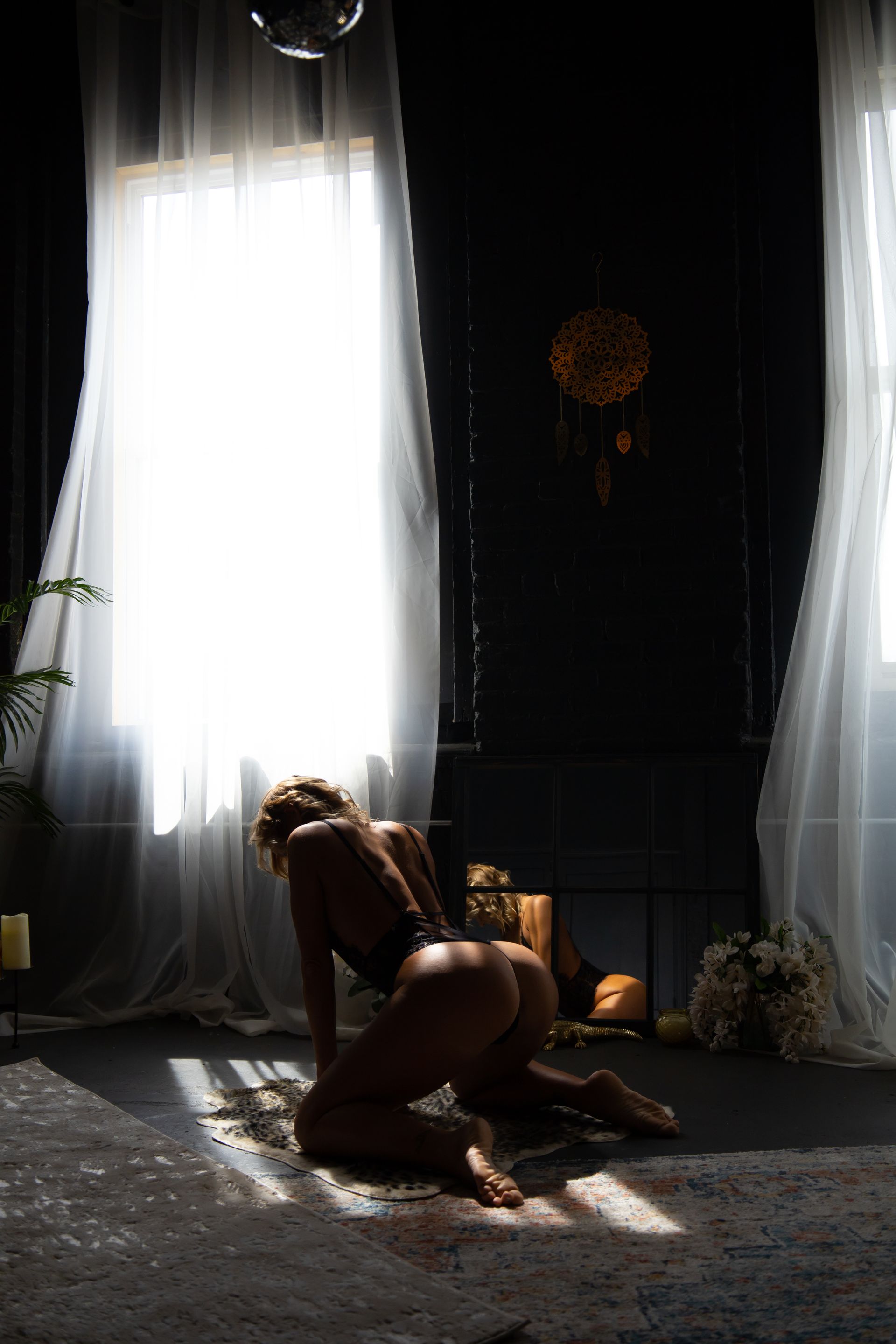 Woman on knees, bending over with back to the camera, near a window with white curtains and a dark wall.