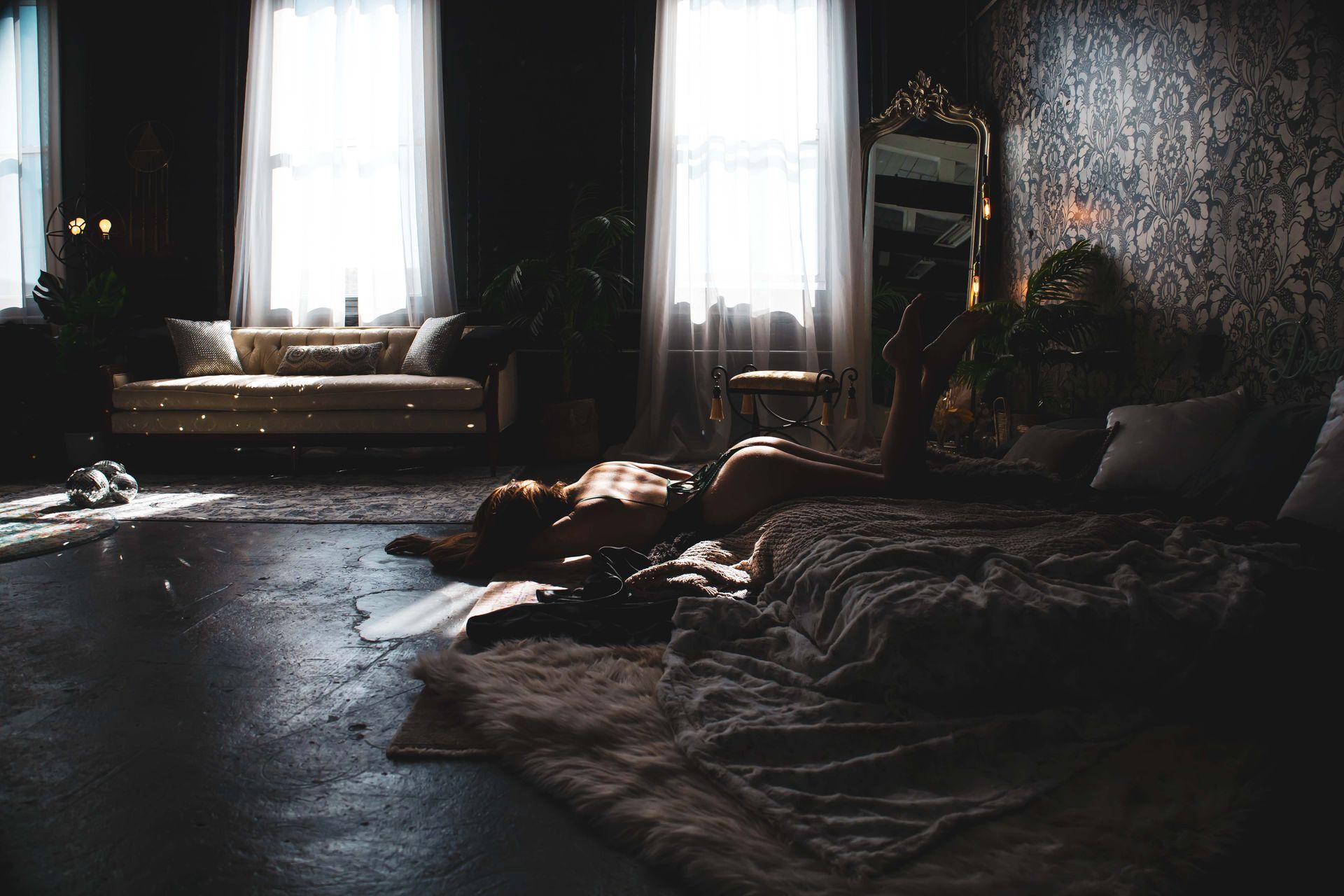 A dimly lit bedroom. A person lies on a bed with a faux fur throw, and light streams through windows onto the floor and couch.