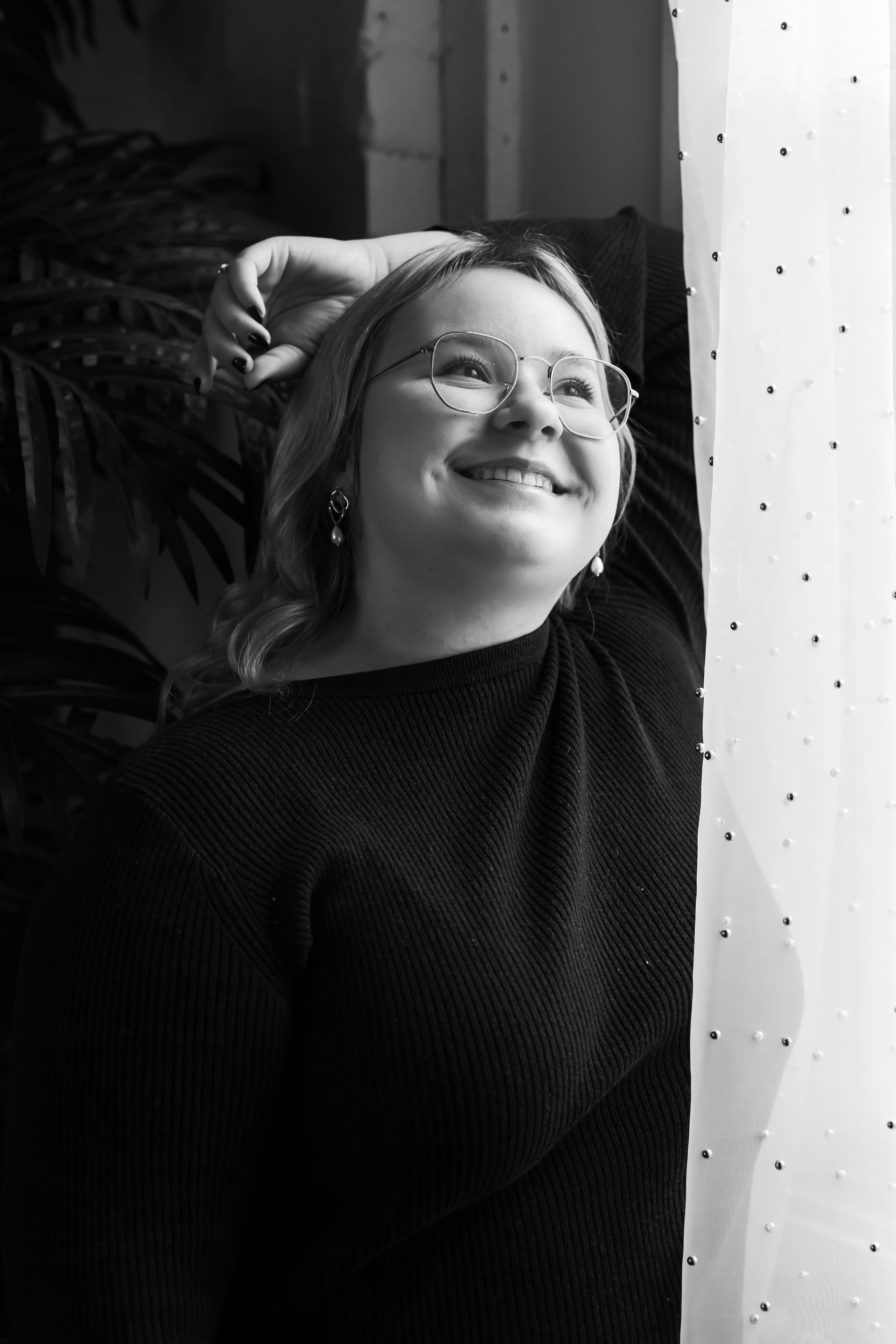Woman with glasses smiles and looks upward, near a window; black sweater, hand on head.
