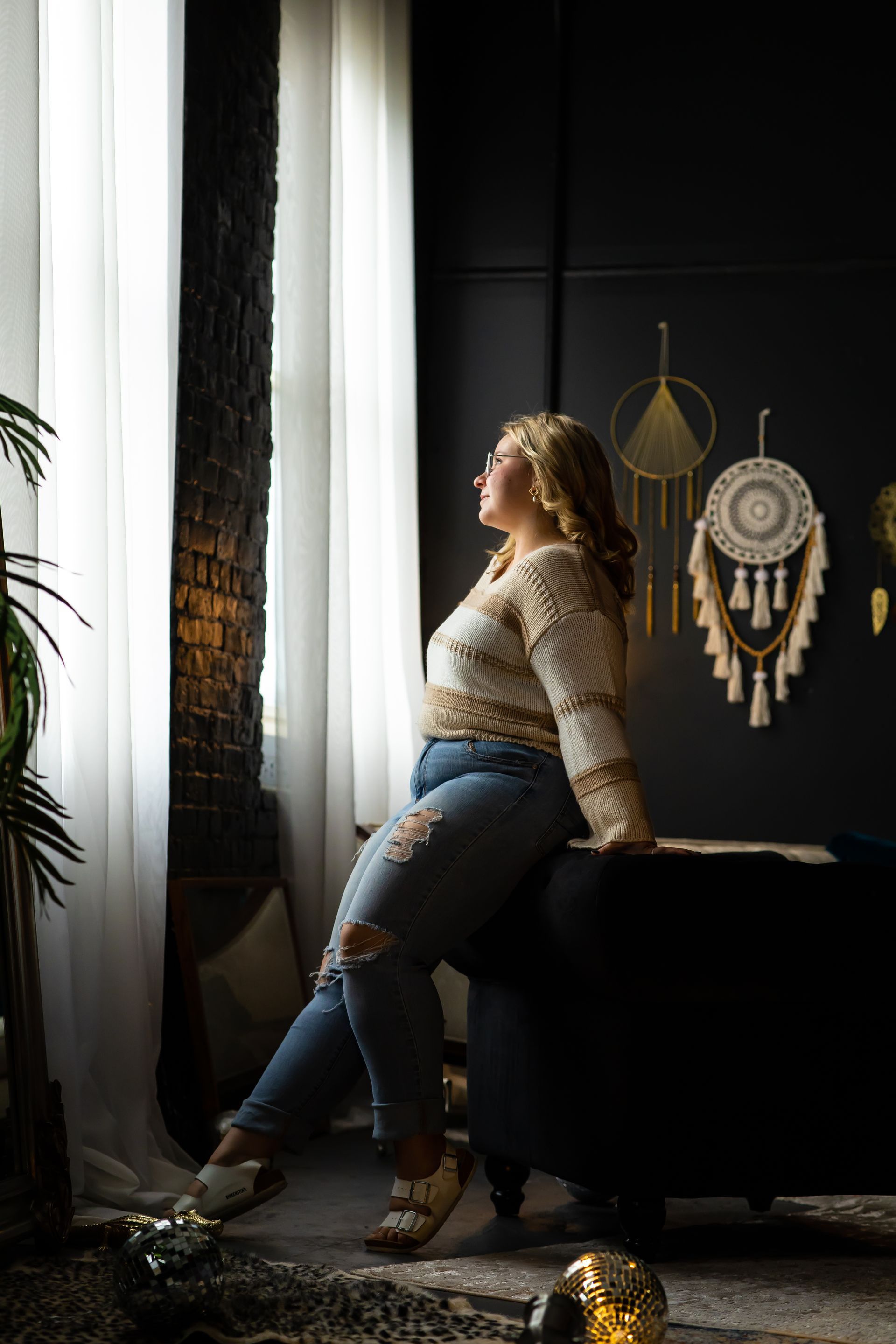Woman in ripped jeans and crocheted sweater leans against a dark couch, looking out a window. Sunlight streams in, illuminating her and the room.