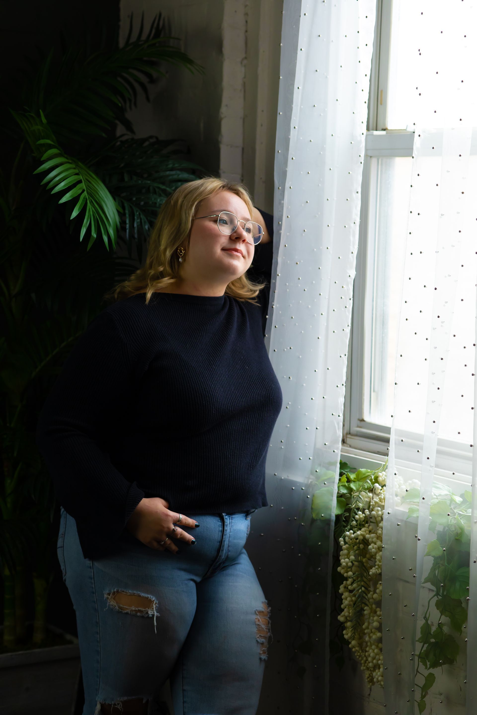 Woman with blonde hair wearing glasses, blue sweater, and ripped jeans, looking out a window.