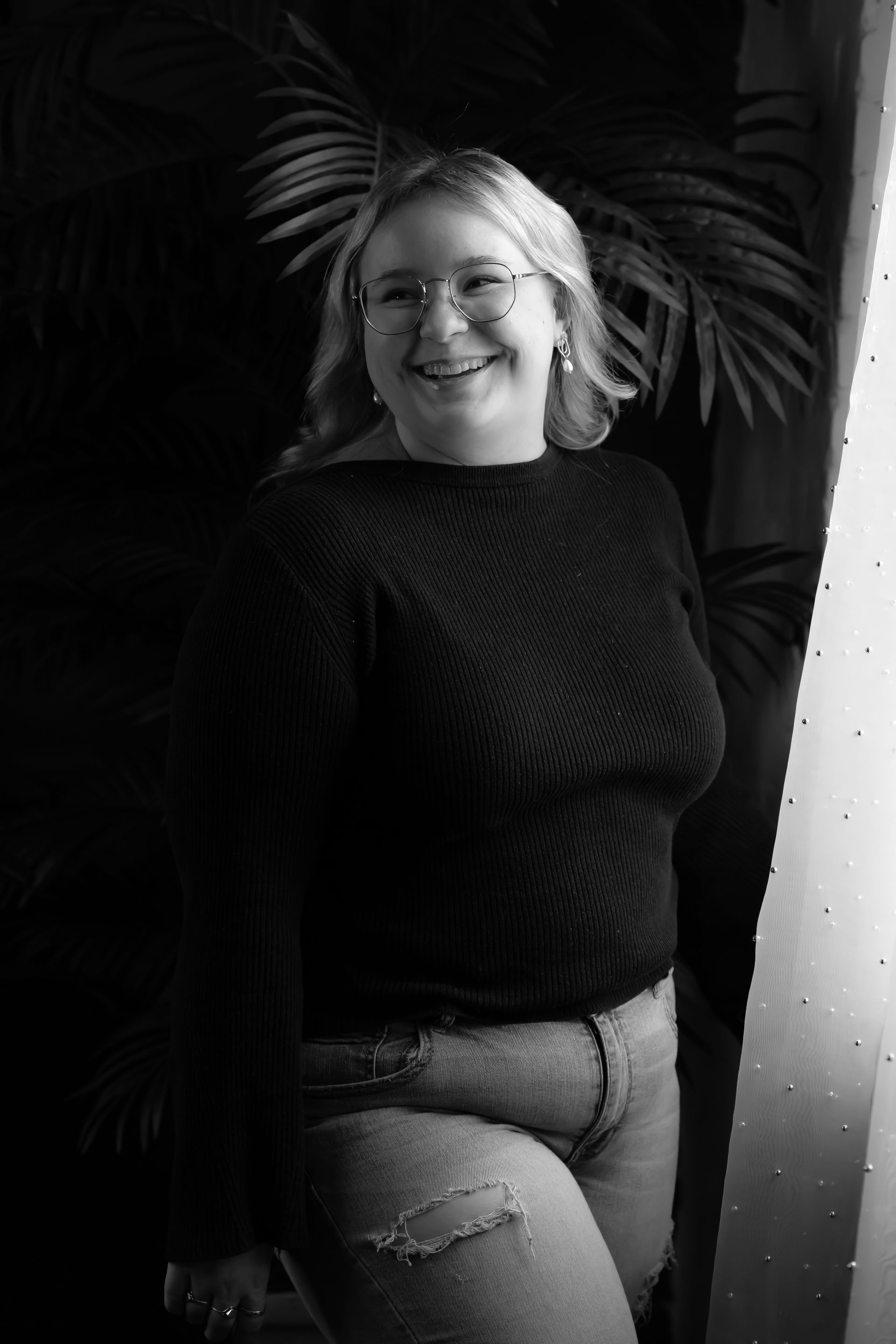 Woman with blonde hair, glasses, and a black sweater smiles, leaning against a wall.