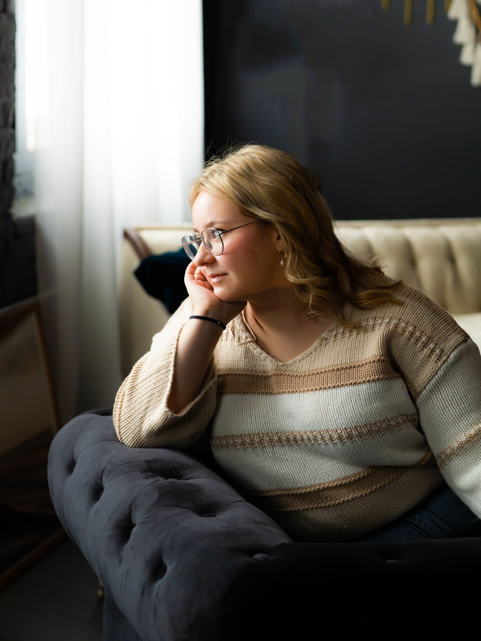 A young woman with glasses rests her chin on her hand, looking out a window. She sits on a dark blue couch, wearing a beige sweater.