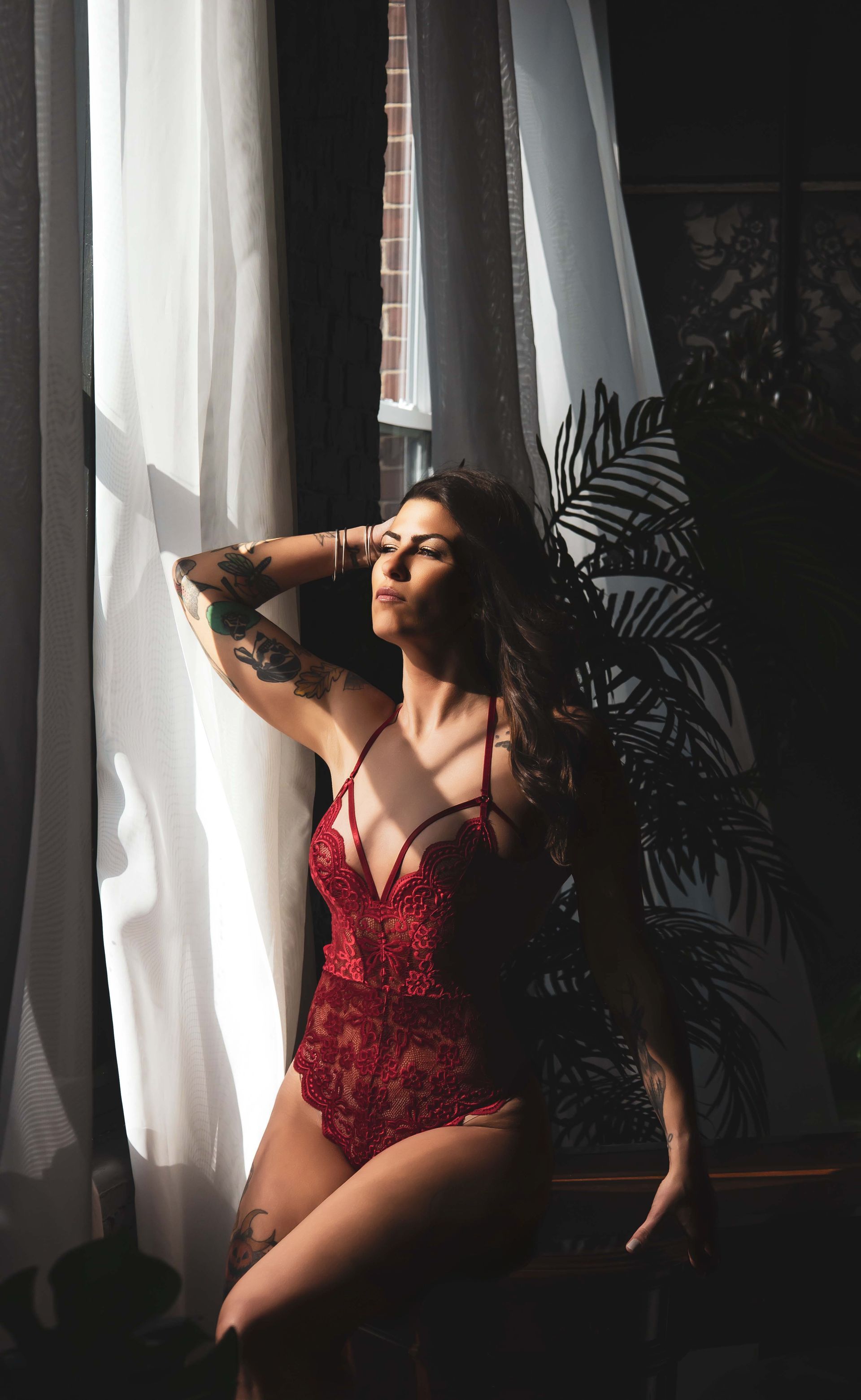 Woman in red lingerie poses by a window with sunlight streaming through sheer curtains. She has tattoos and long hair.