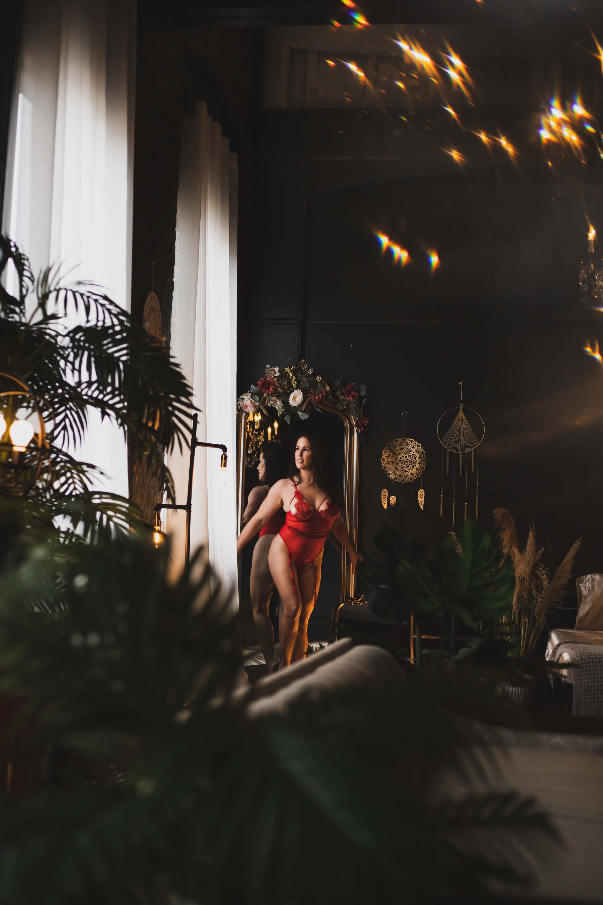 Woman in red lingerie stands near a tall window in a dark room with lush plants. Light sparkles.