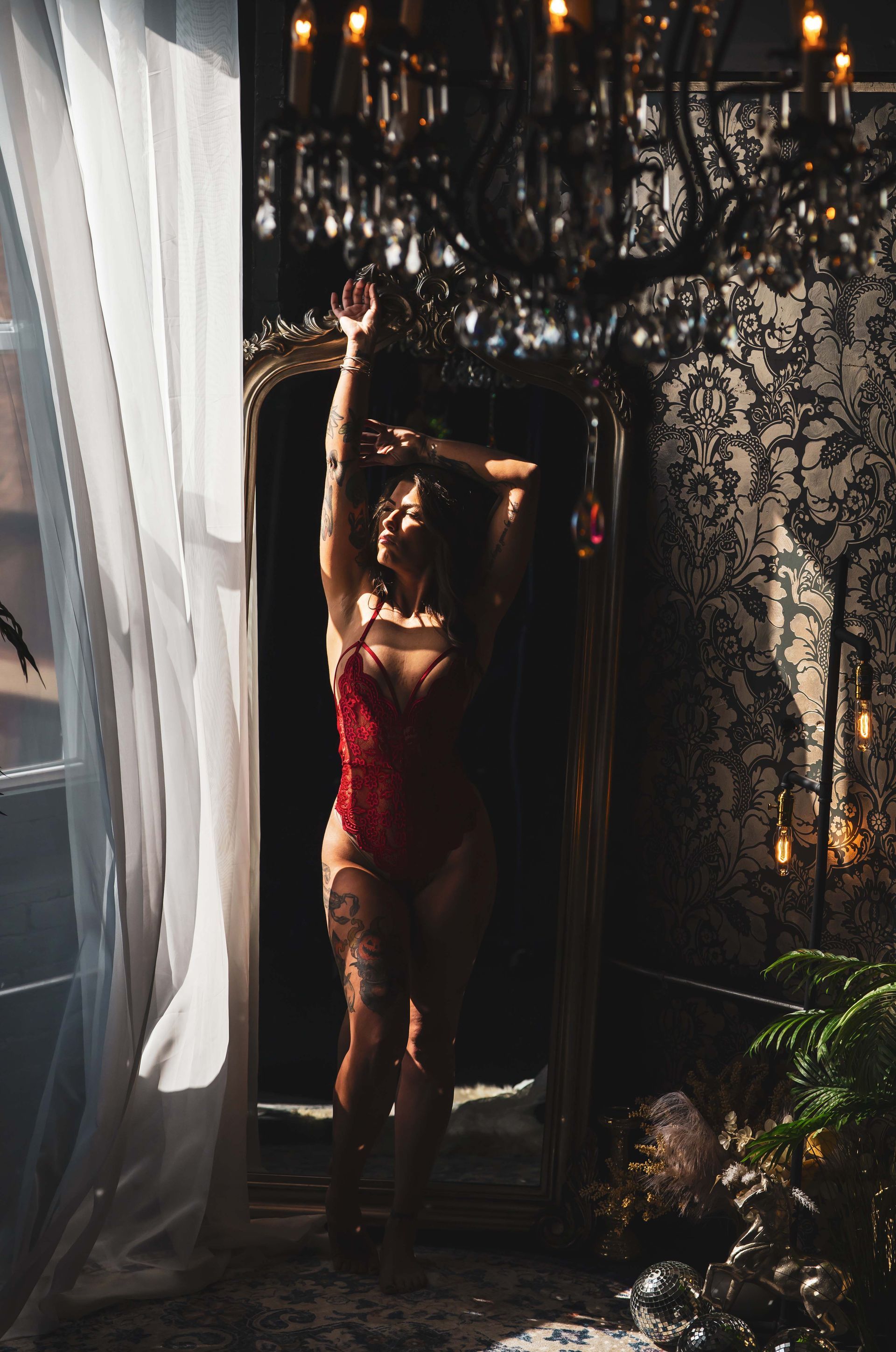 Woman in red lingerie poses in front of an ornate mirror, arms raised. Bright sunlight streams through a window; elegant room.