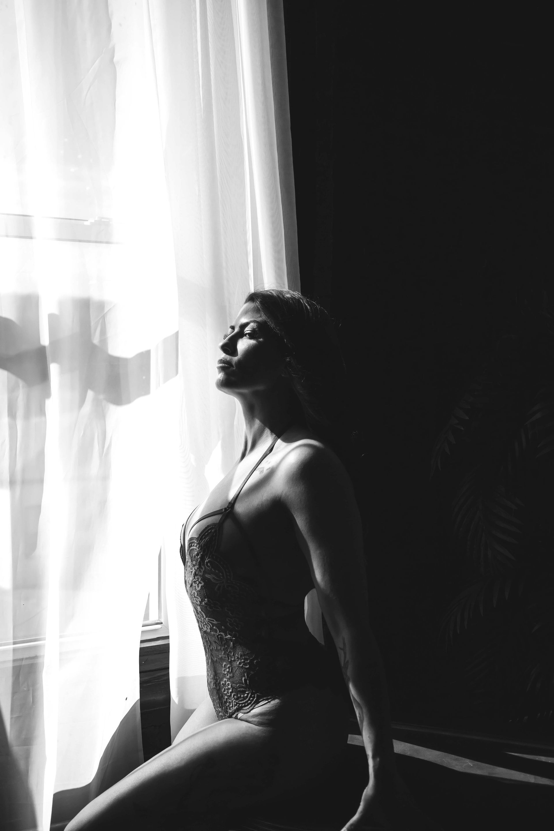 Woman in lingerie, backlit by a window, tilting her head up in a contemplative pose. Black and white.