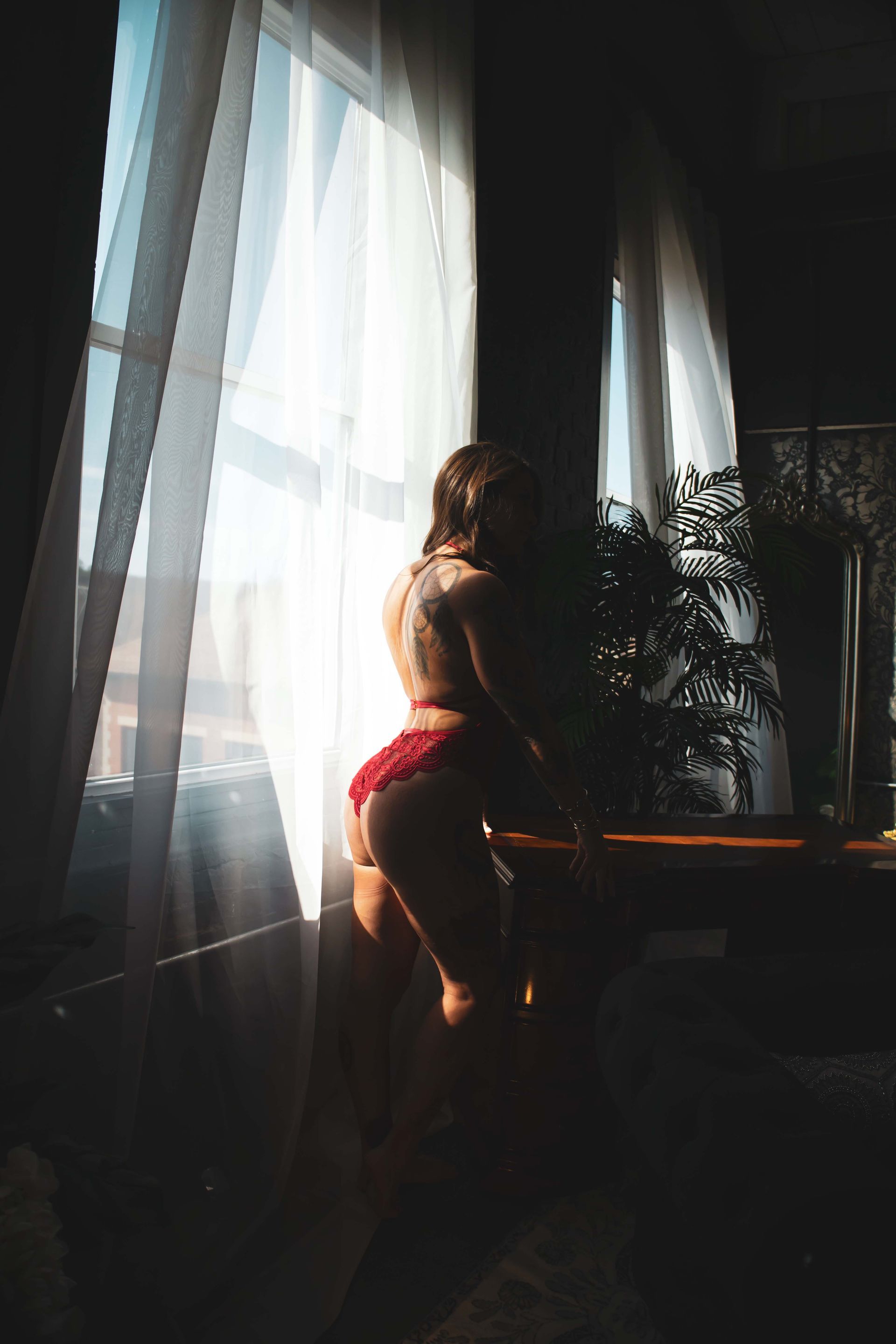 Woman in red lingerie poses near a window, back to the viewer, with sunlight streaming in.