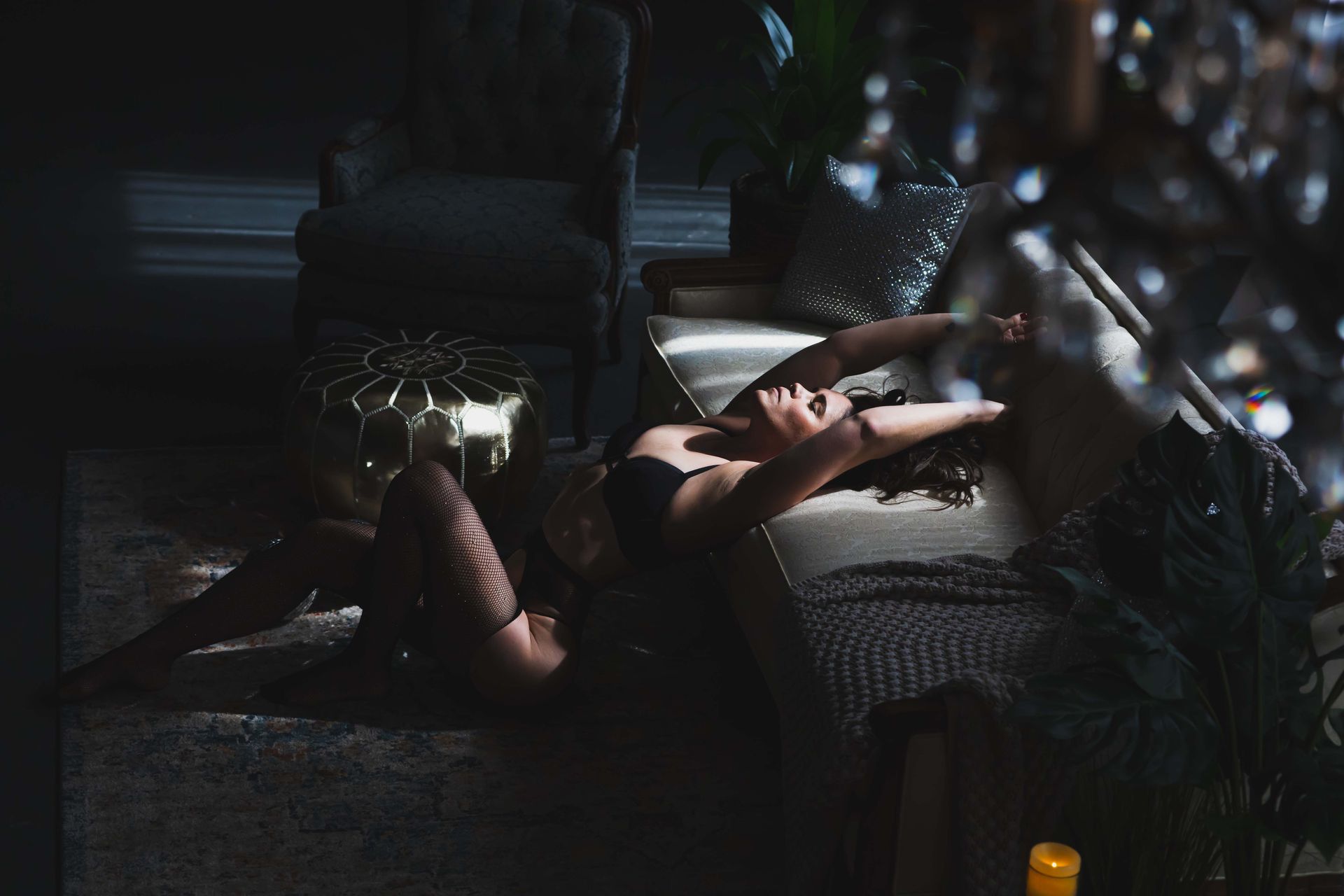 Woman in lingerie reclines on a couch, lit by sunlight. Dark hair, black bra, and shadows create a sensual atmosphere.