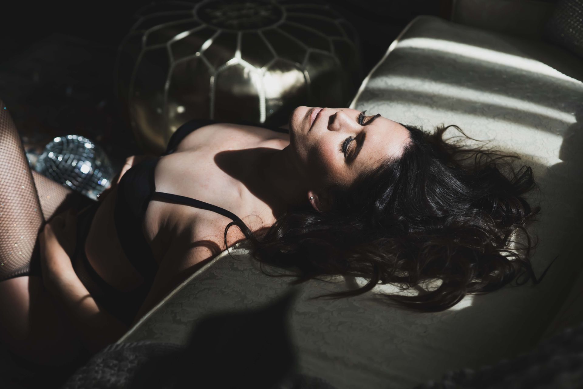 Woman lying on a couch in lingerie, bathed in sunlight. Dark hair, eyes closed, neutral expression.