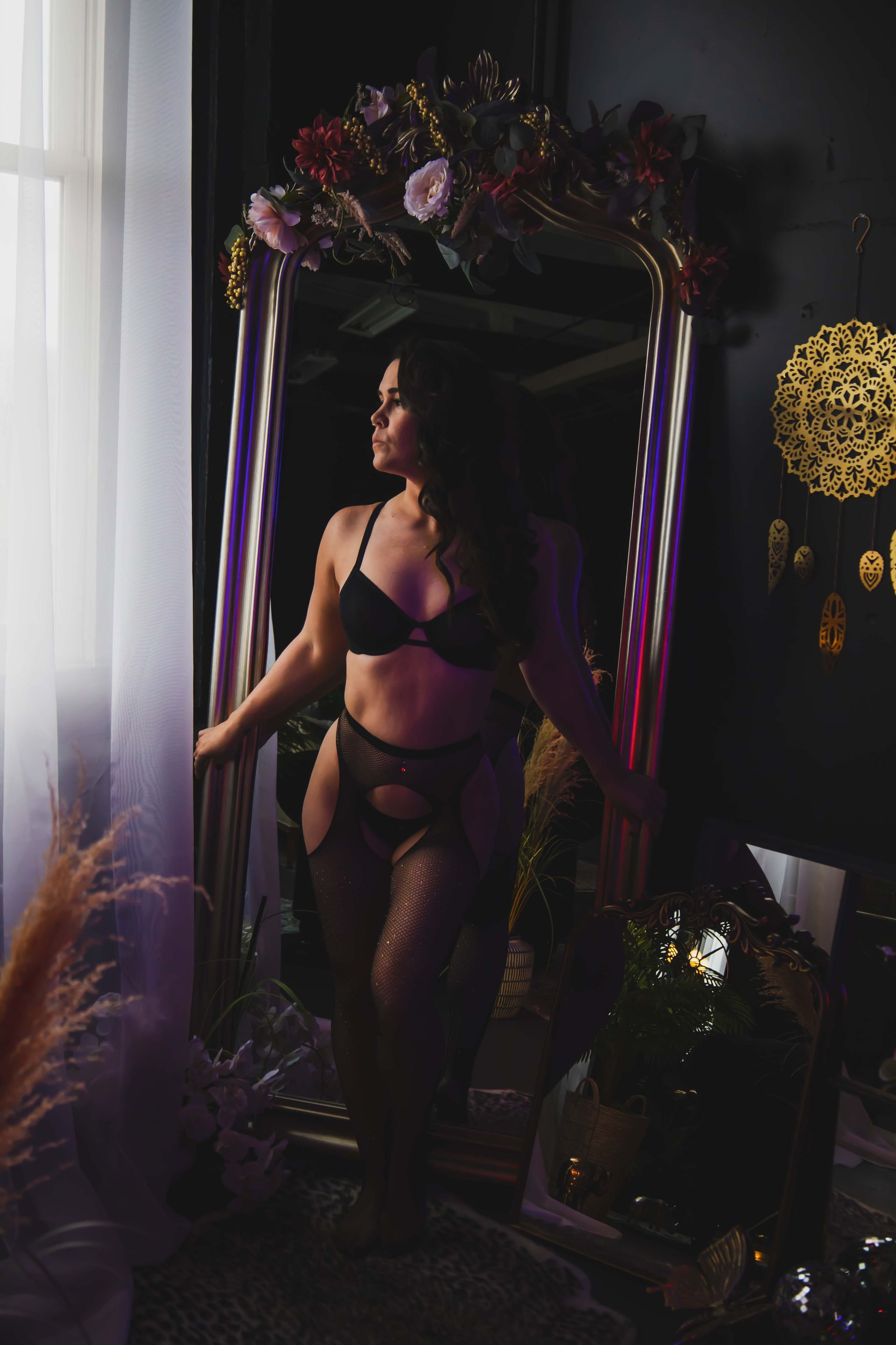 Woman in black lingerie poses in front of a large mirror, looking towards a window. The mirror is adorned with flowers.