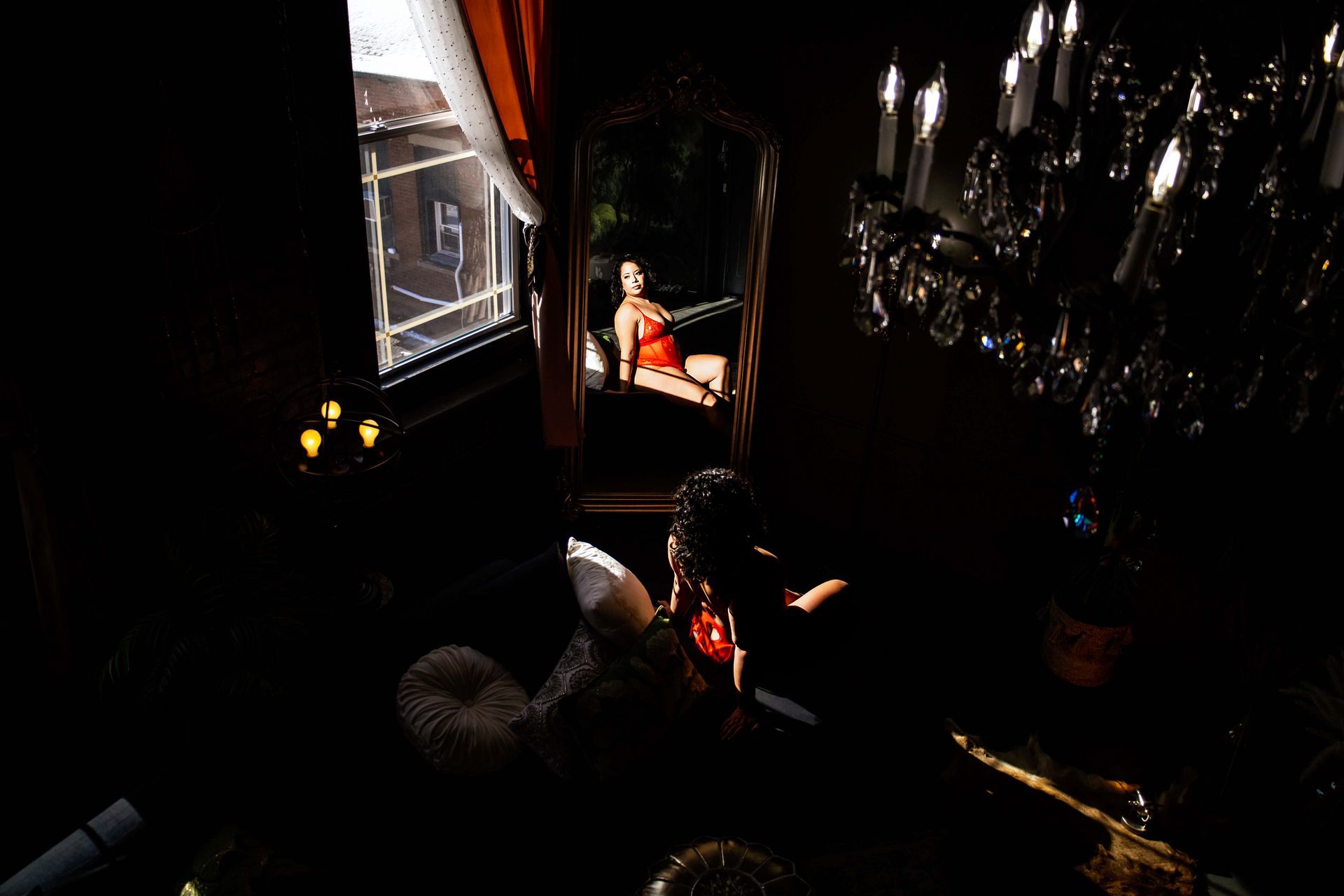Mirror reflecting woman in red dress, room with ornate chandelier, candles, and window.