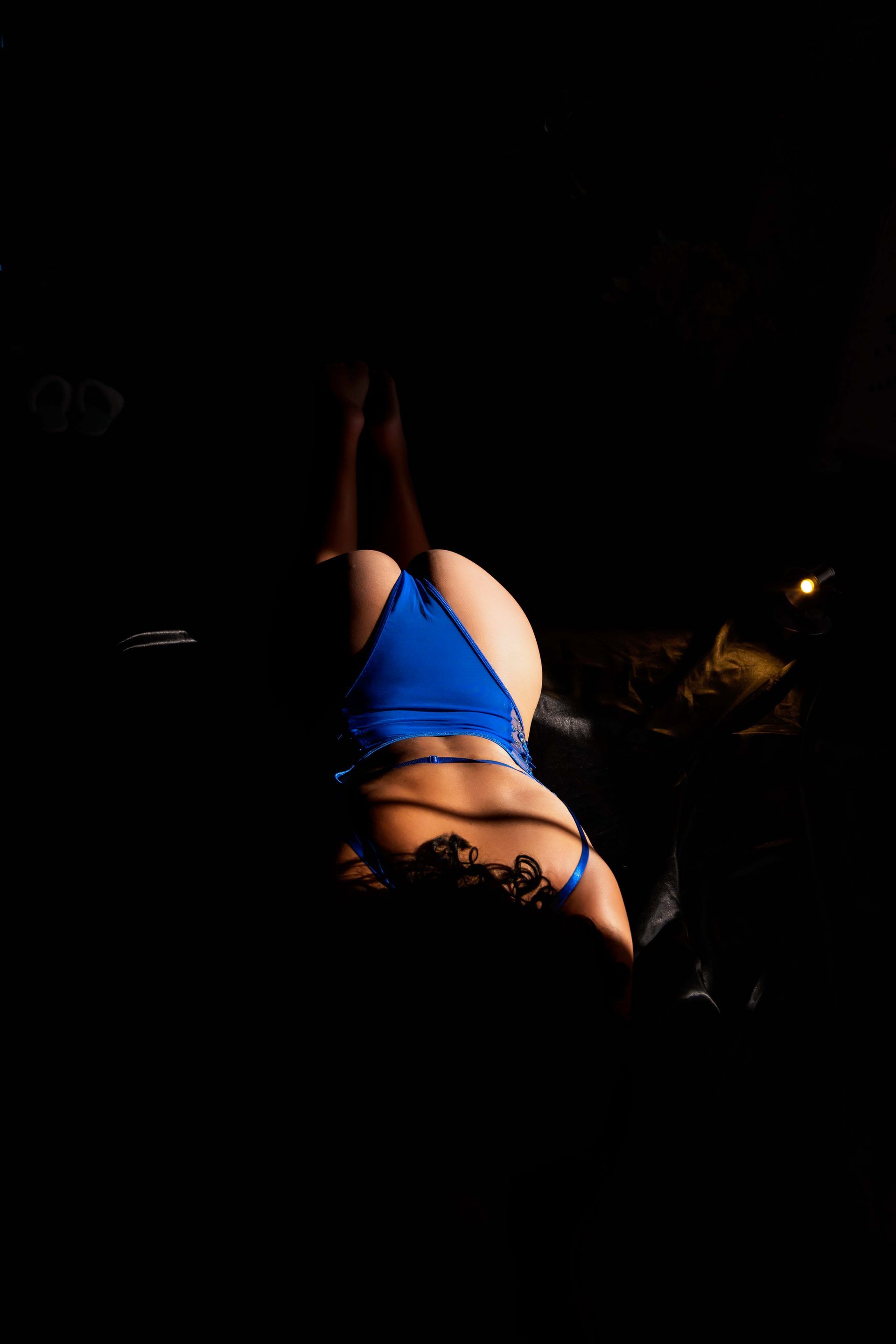 Woman in blue swimsuit, lying on stomach with legs raised in the dark.