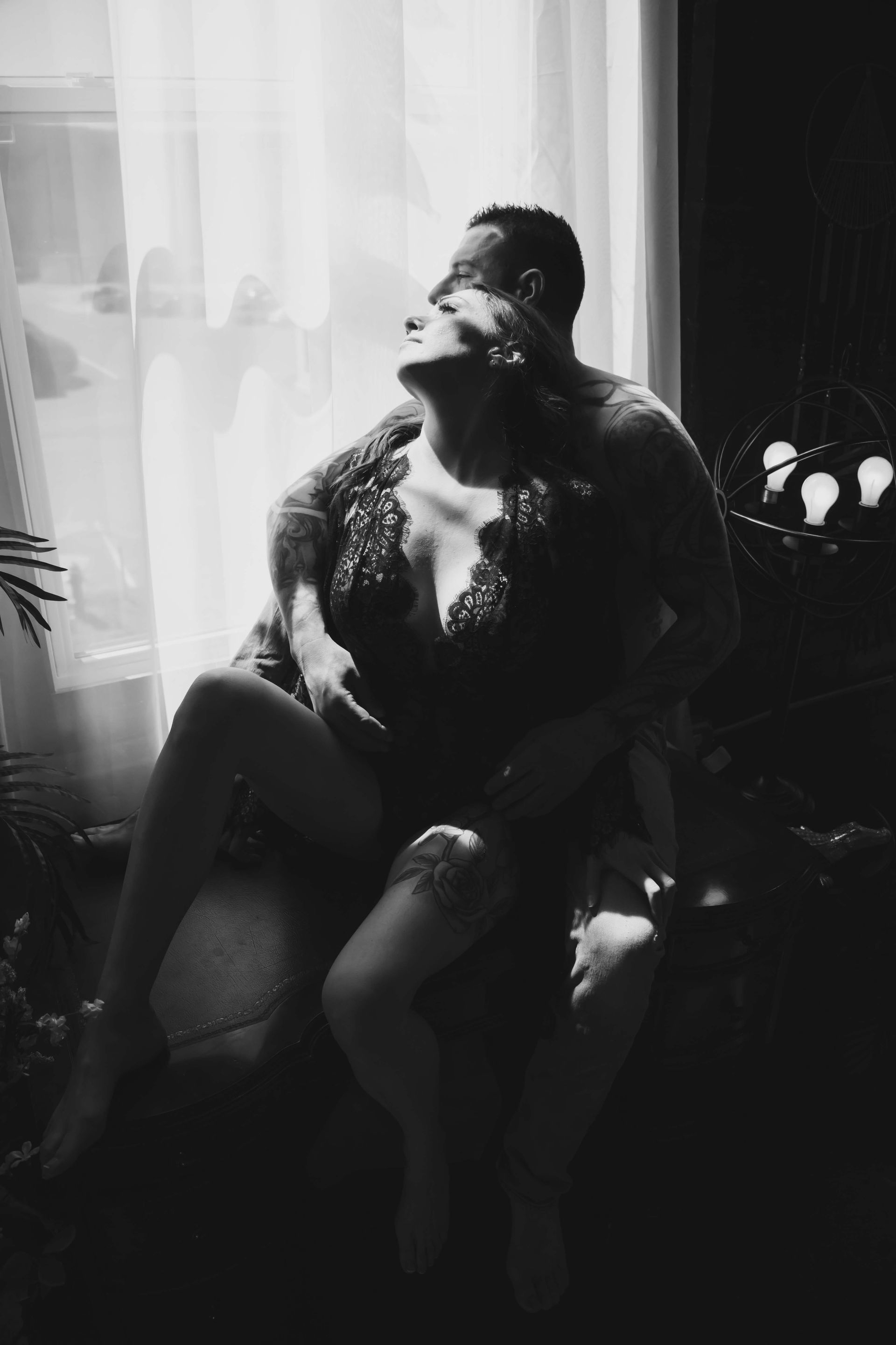 A couple embracing in front of a window. The woman sits on the man's lap, both partially undressed in a dimly lit room.