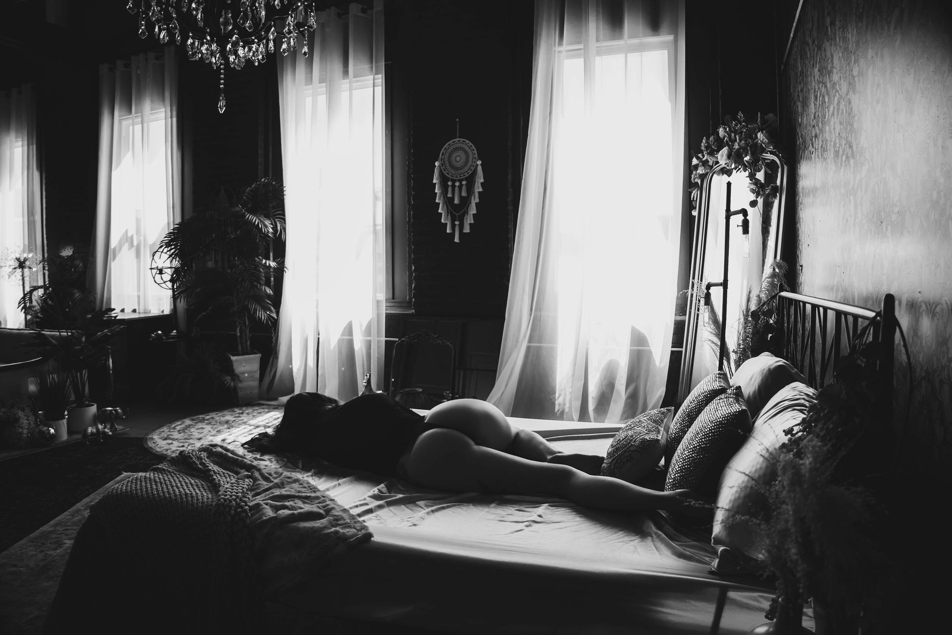 Black and white shot of a person lying on a bed in a room with large windows and curtains. A chandelier hangs overhead.