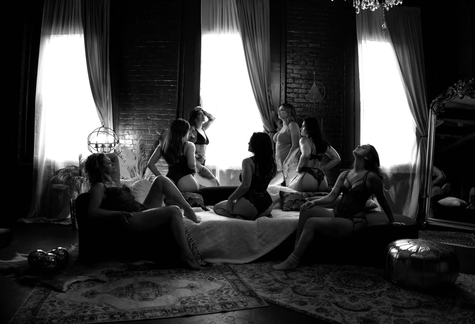 Backlit room with several people lounging and sitting on a bed near tall curtained windows
