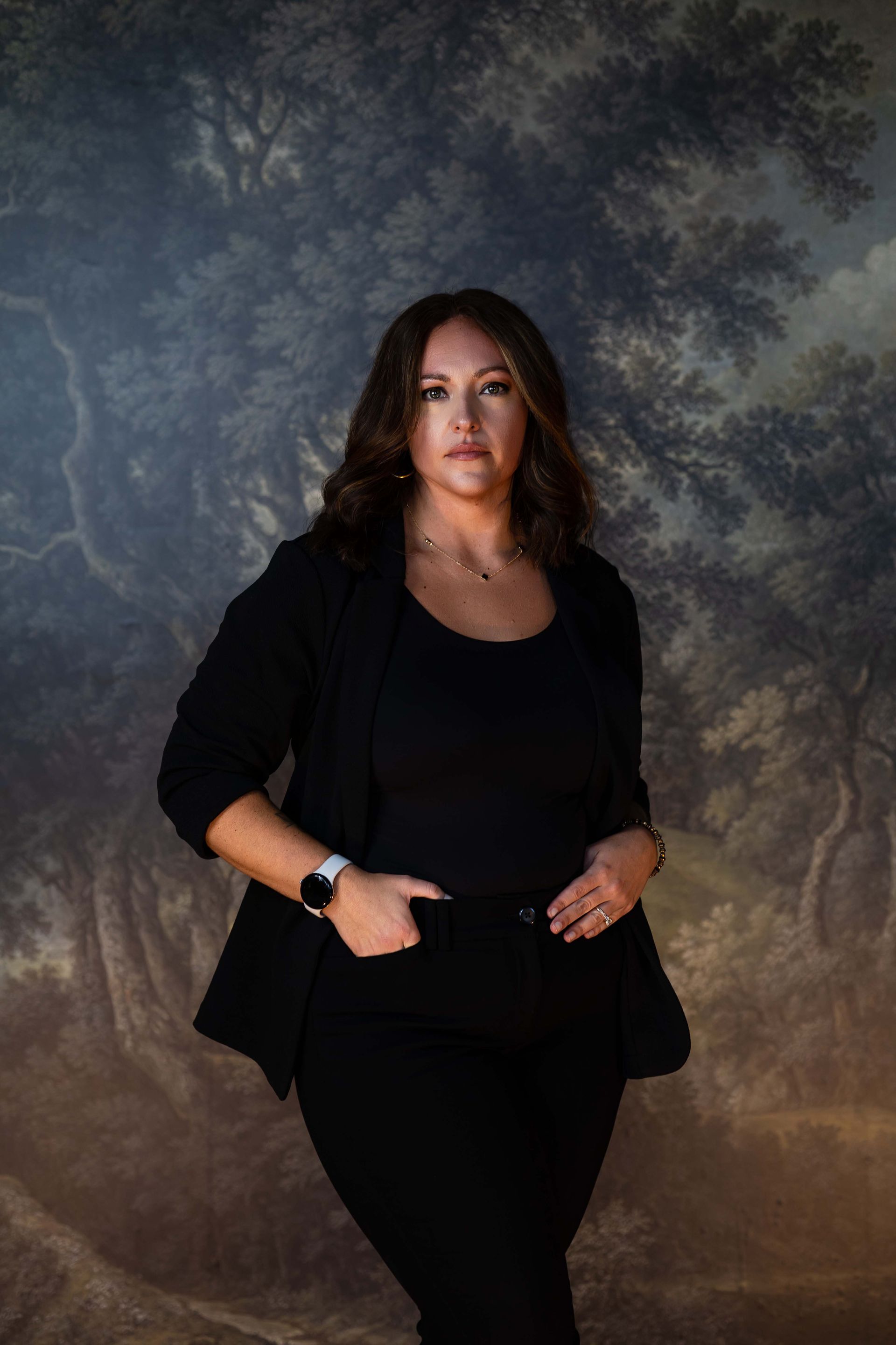 Woman in a black outfit standing with hands in pockets against a dark textured backdrop.