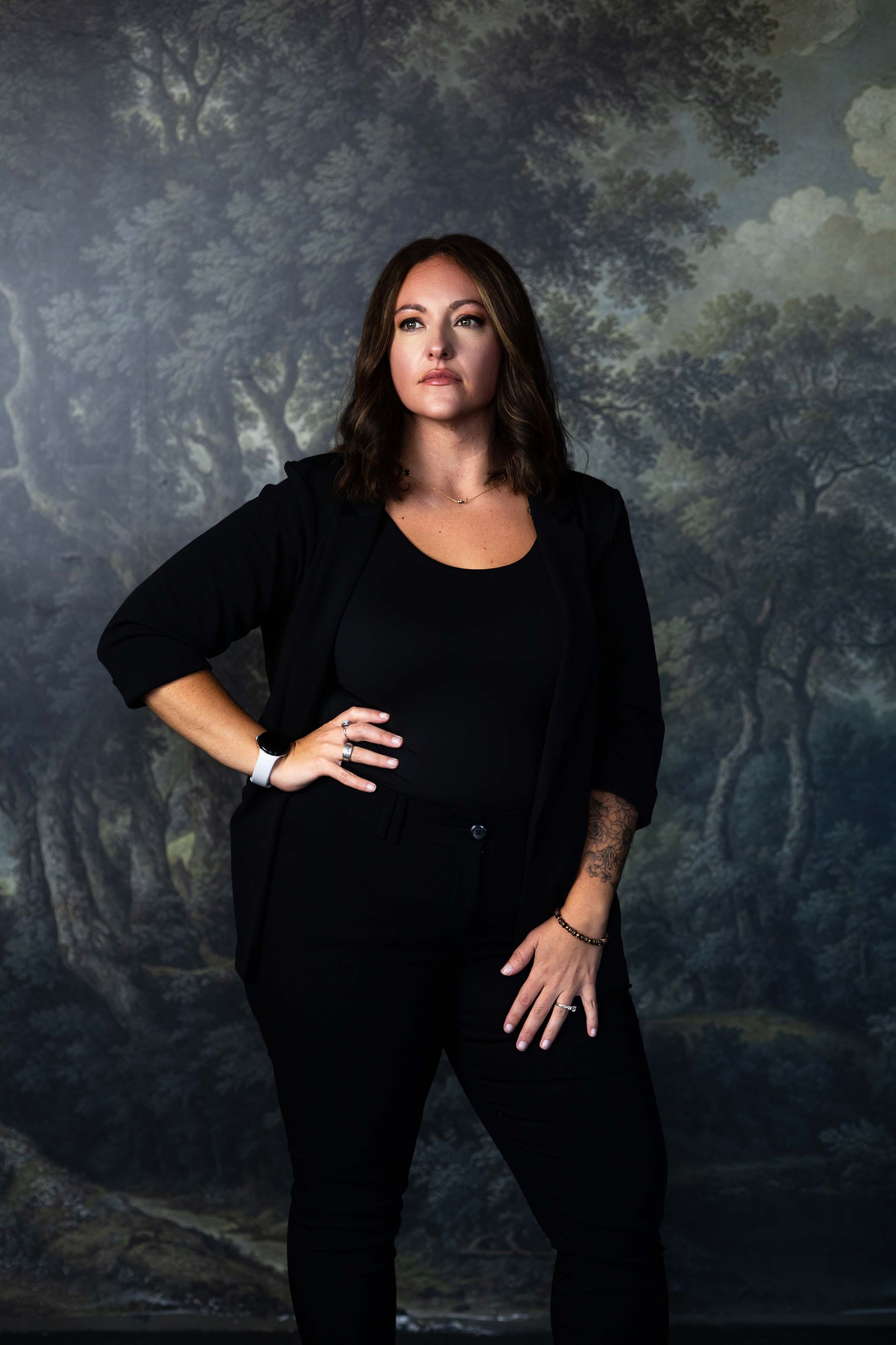 Woman in black posing against a dark forest backdrop, one hand on hip and one at her side.