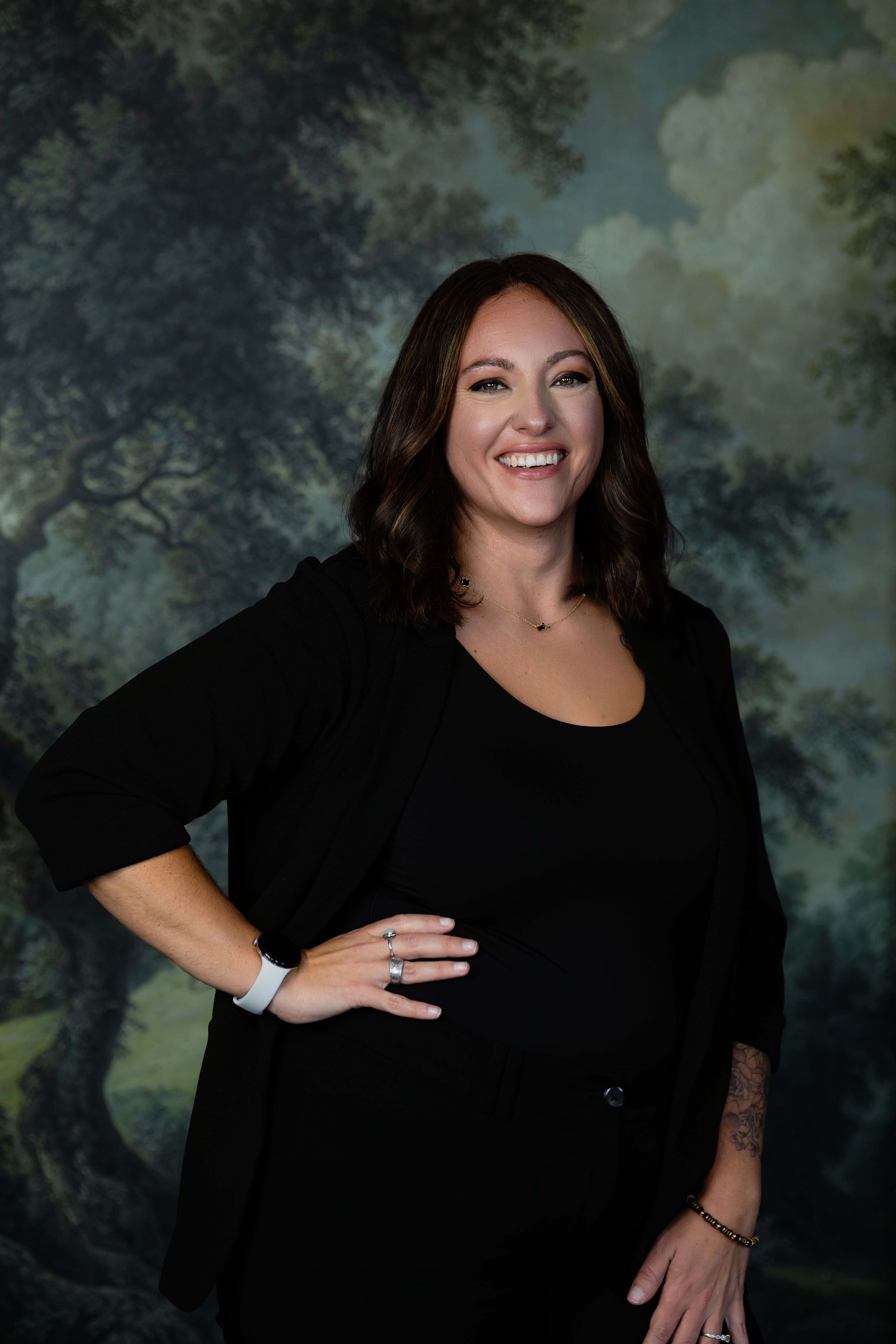 Woman smiling in black outfit, standing with hand on hip against a cloudy studio backdrop