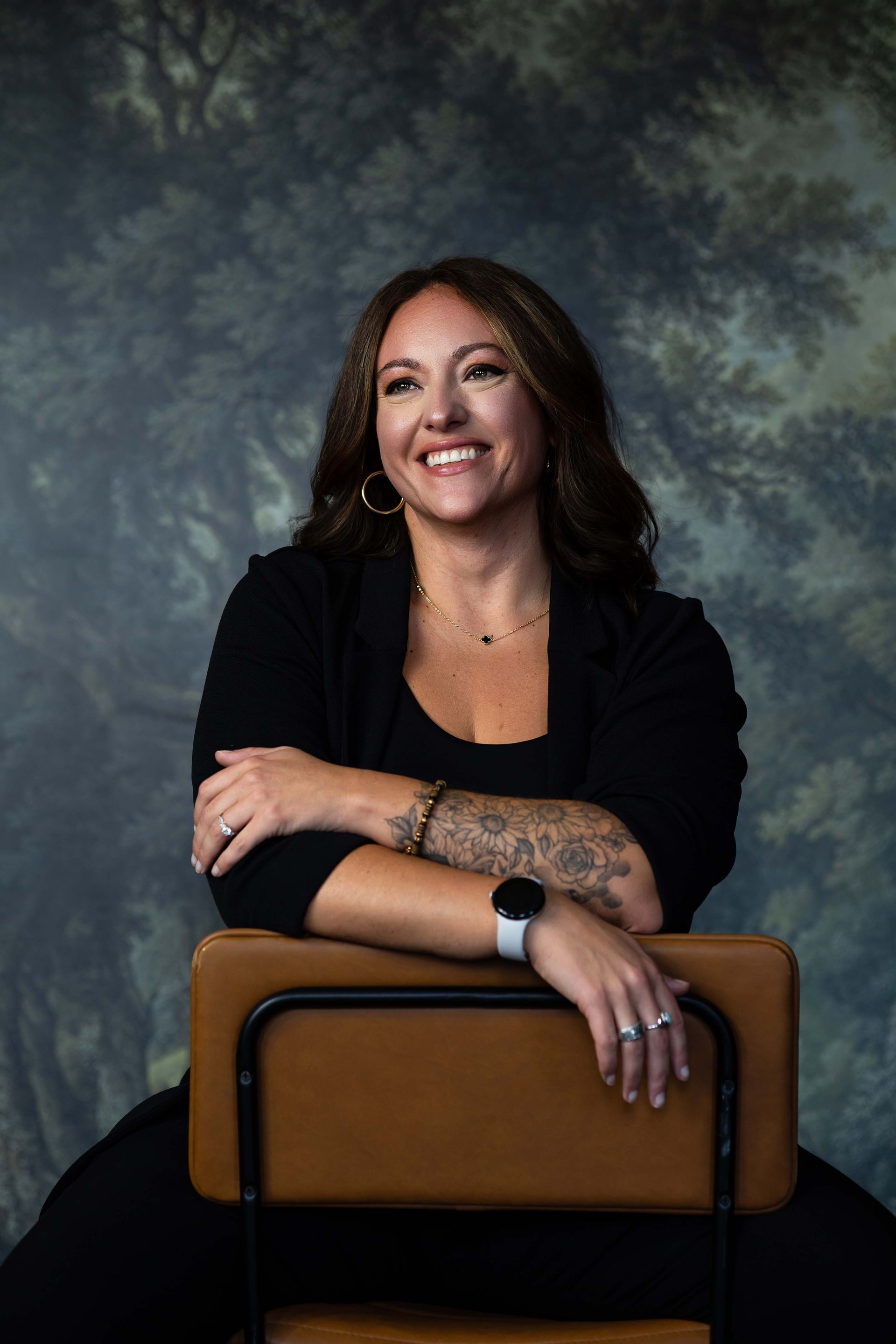 Smiling person leaning on a chair, wearing a black outfit against a mottled studio backdrop.