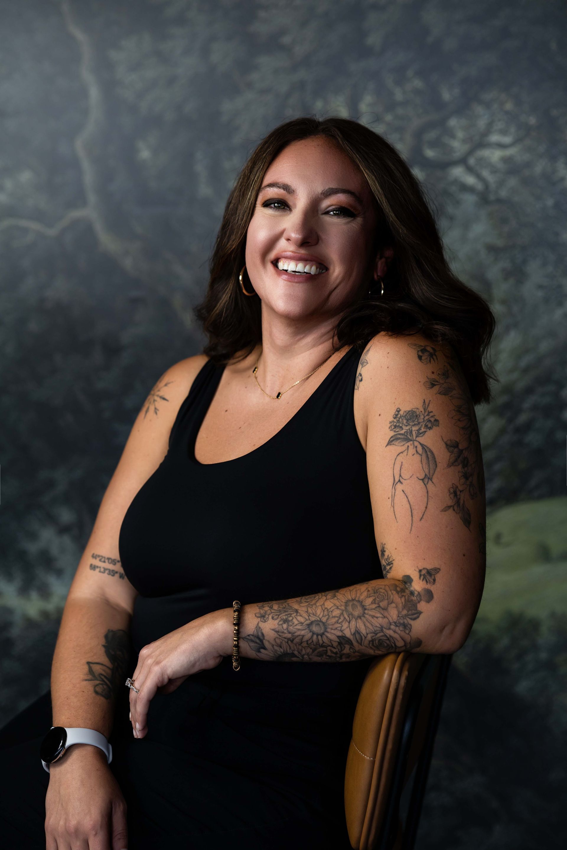 Smiling woman in a black sleeveless top seated against a gray textured backdrop, with tattooed arms.