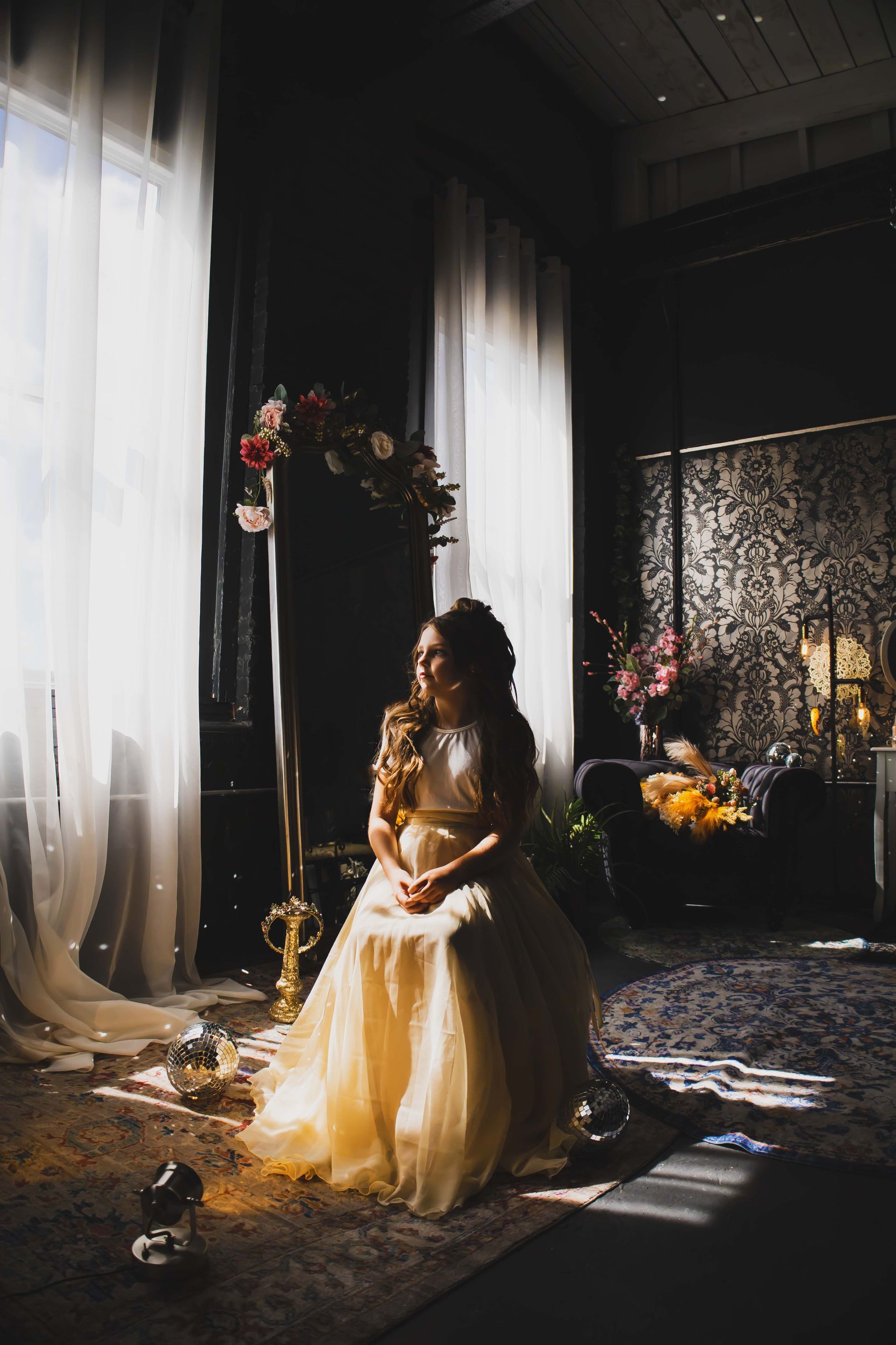 Woman in a cream-colored gown sits near a window with sunlight streaming in. She's in a dimly lit, elegant room with floral arrangements.