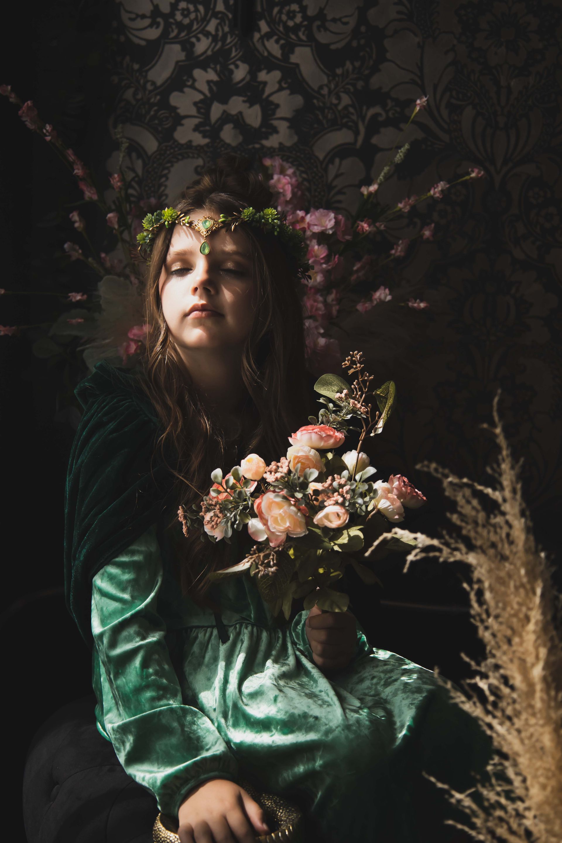 A young person in a teal dress and crown holds flowers, eyes closed, in a dimly lit room with floral wallpaper.