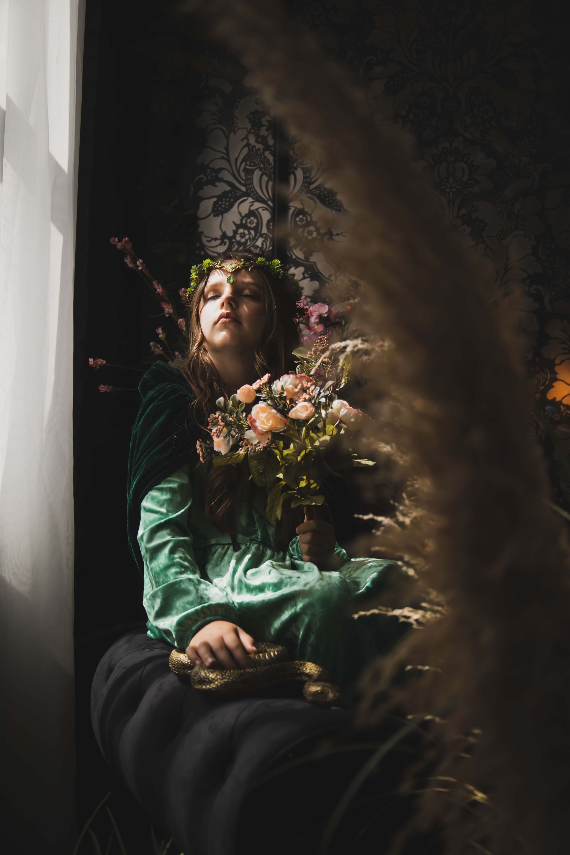 A person in a green dress with flowers and a crown sits near a window, appearing pensive. Soft lighting illuminates the scene with dark and warm tones.