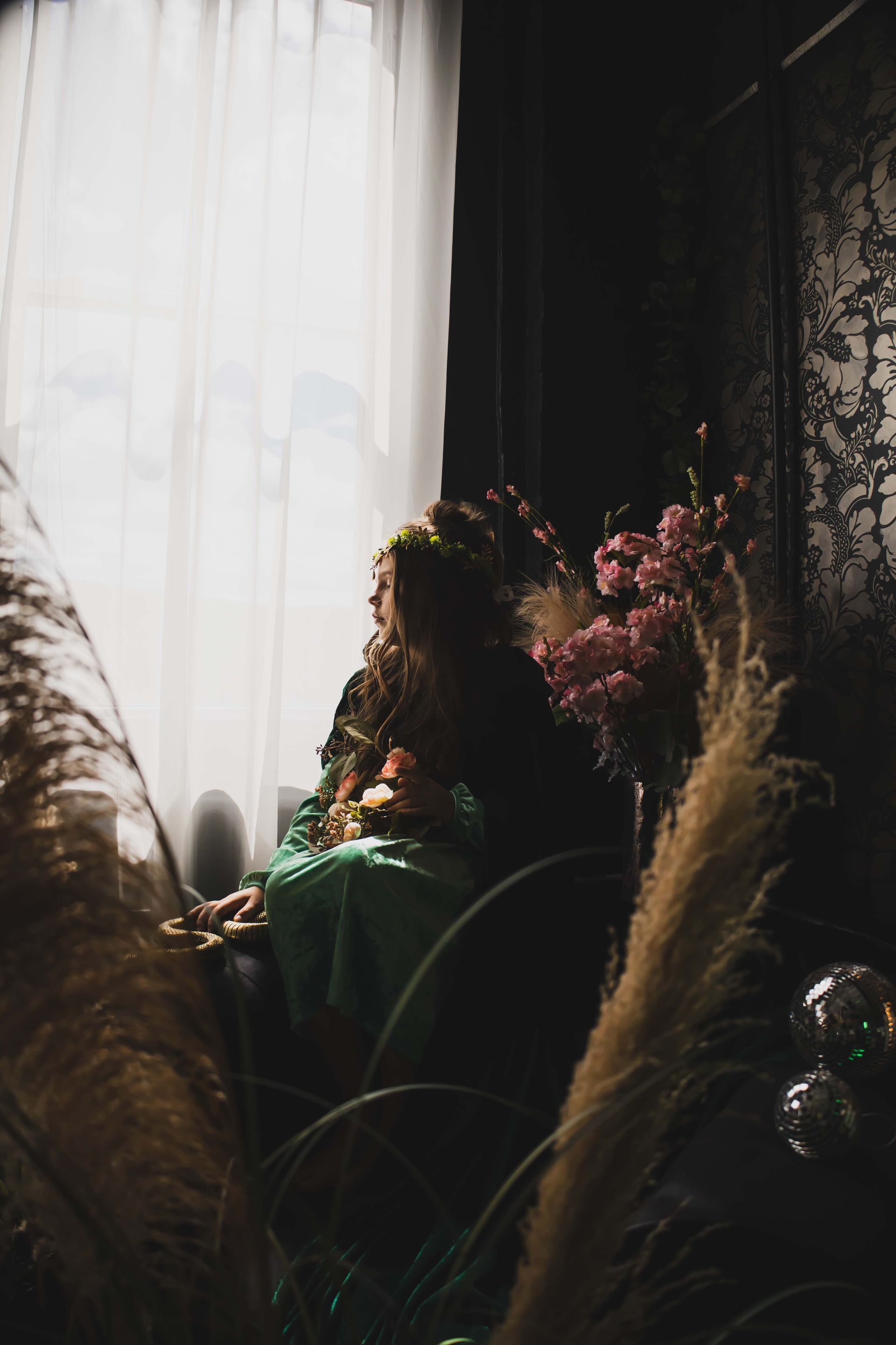 Woman in a green dress looks out a window. Soft light shines on her and a vase of pink flowers.