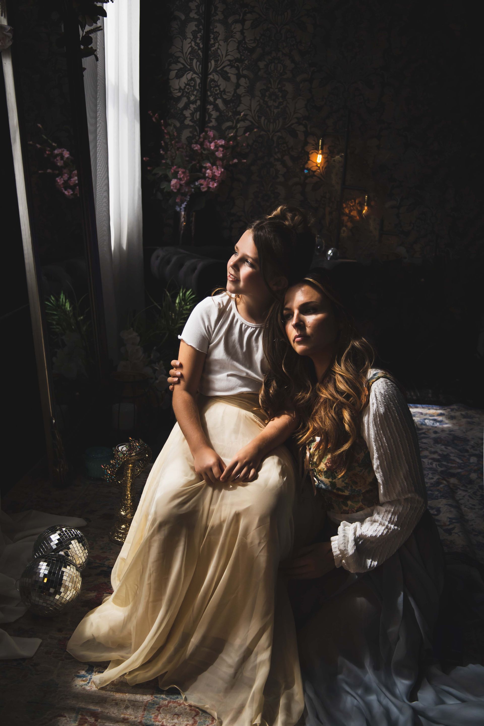 Two females seated in a dark room near a window. One, a child, looks up with an arm around the shoulder of an older woman.