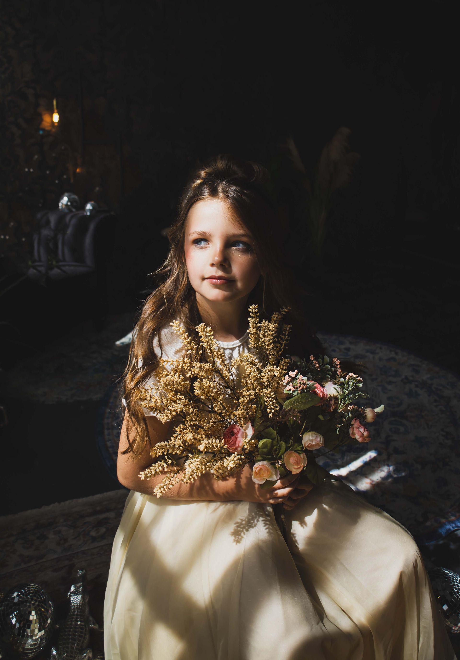 A young girl in a cream dress sits holding a bouquet, illuminated by sunlight filtering through a dark room.