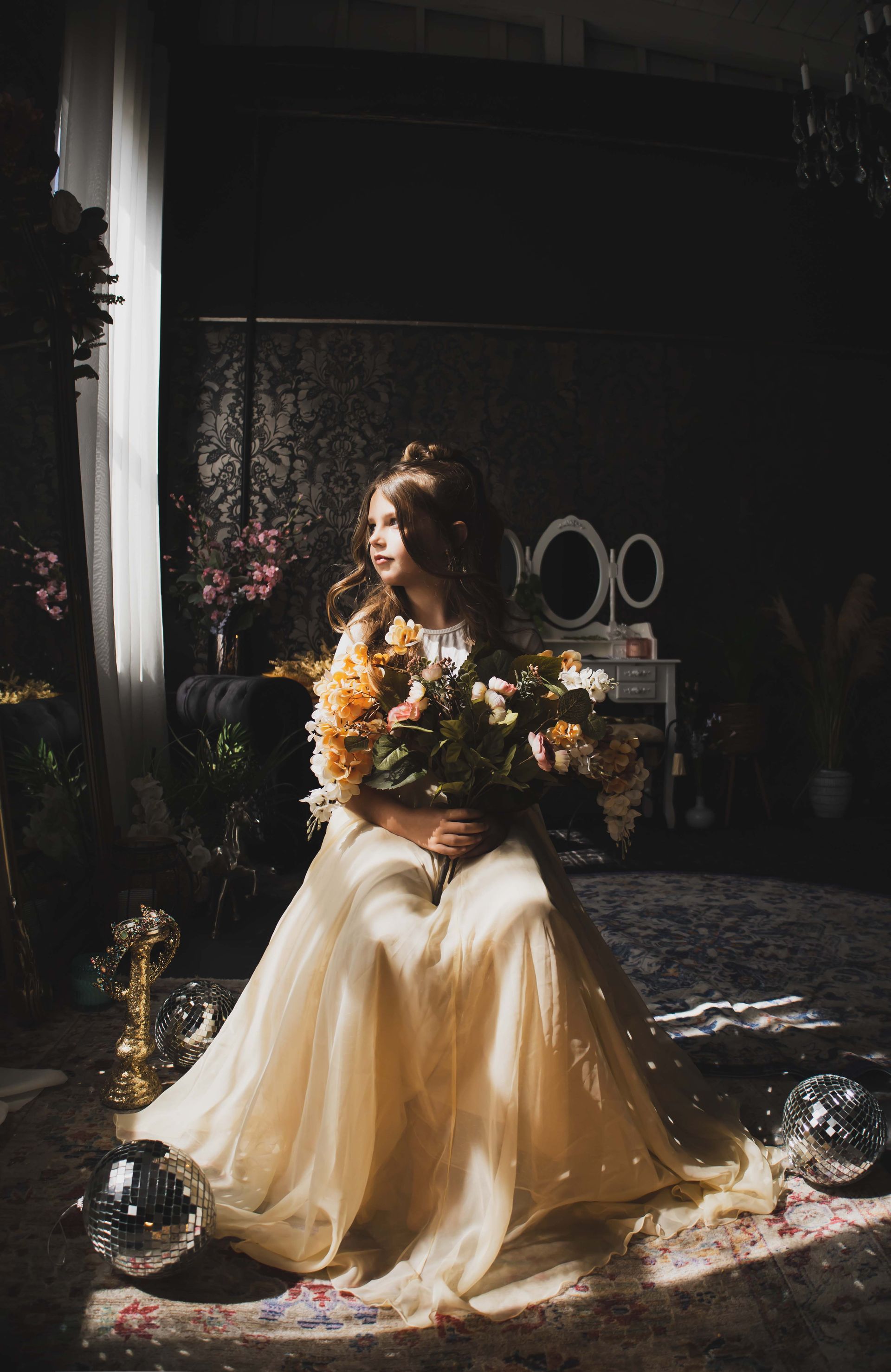 Woman in a flowing gold dress sits in a dark room, holding a bouquet of flowers.  She looks away, lit by a beam of light.