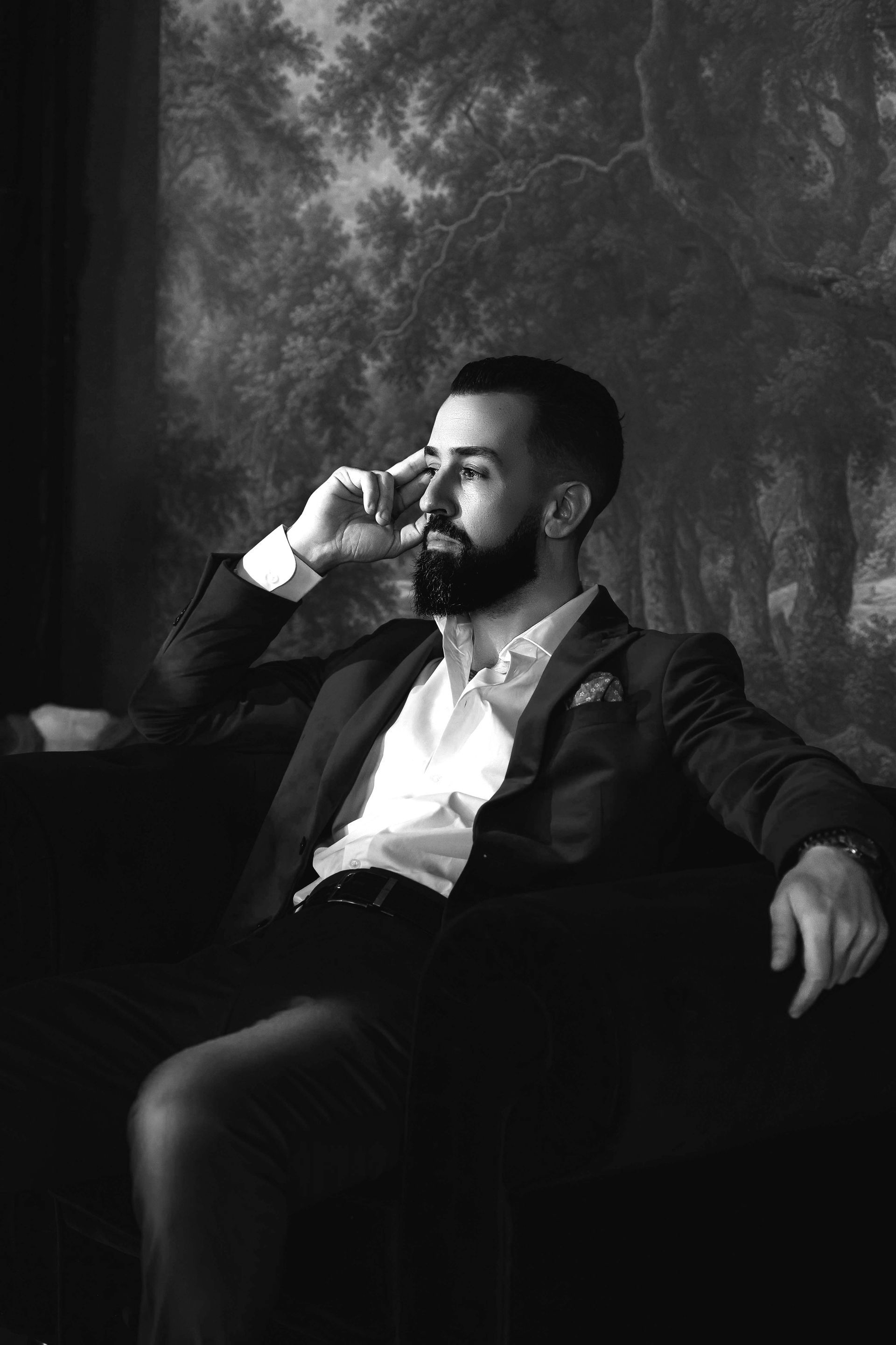 Man in suit, seated, holding phone to ear, looking off-camera. Dark background, thoughtful expression.
