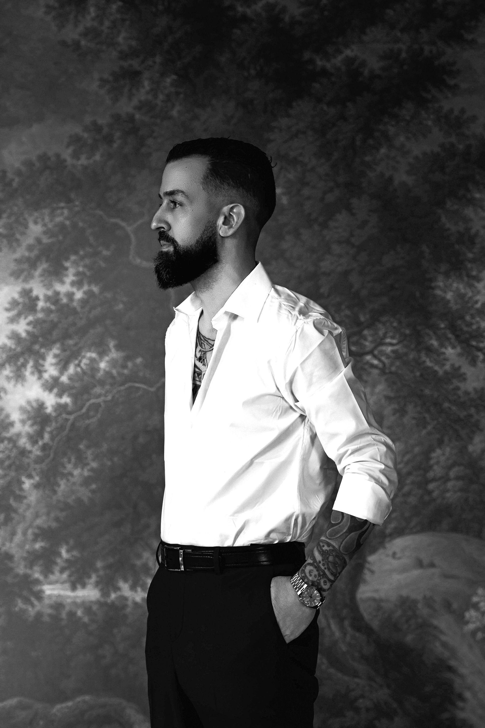 Man in white shirt, black pants, arm tattoos, standing in front of a painted backdrop.