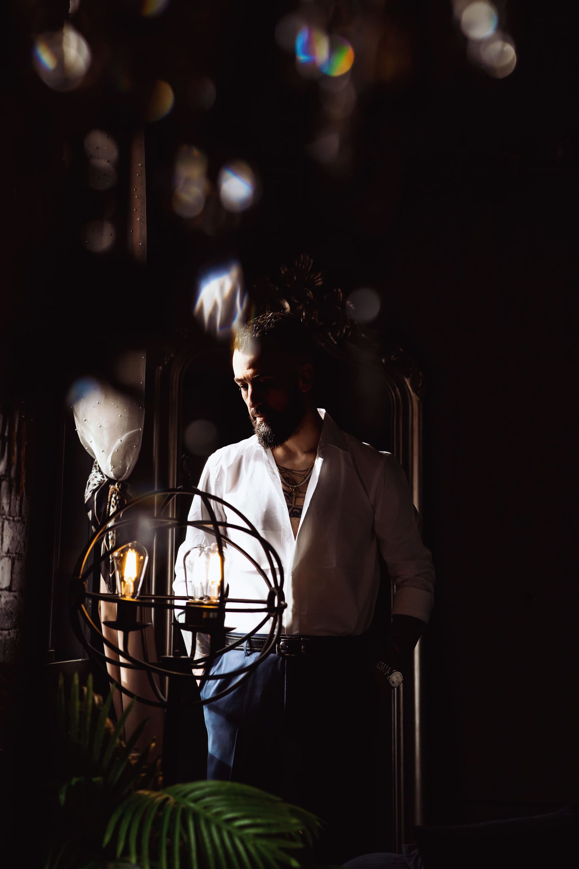 Man in white shirt in dimly lit room, standing near a decorative lamp and plant.