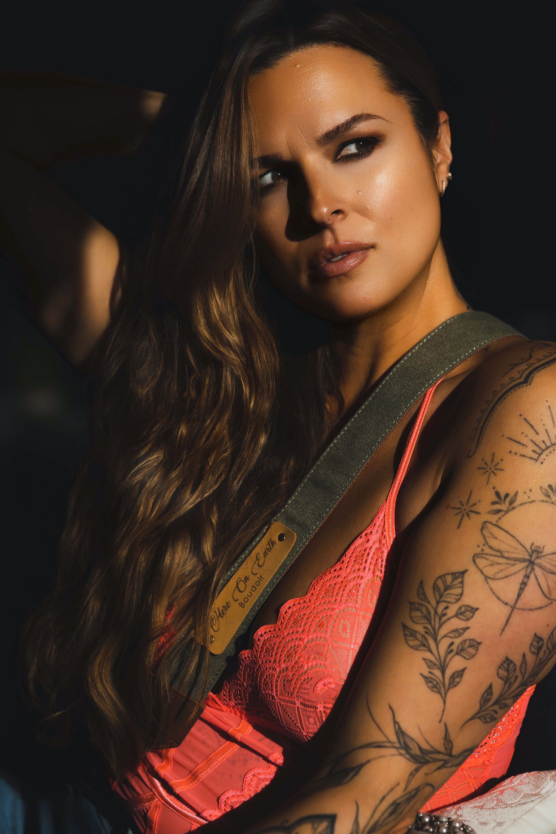 Woman with long brown hair, wearing a pink lace top, with arm tattoos. Dramatic lighting.