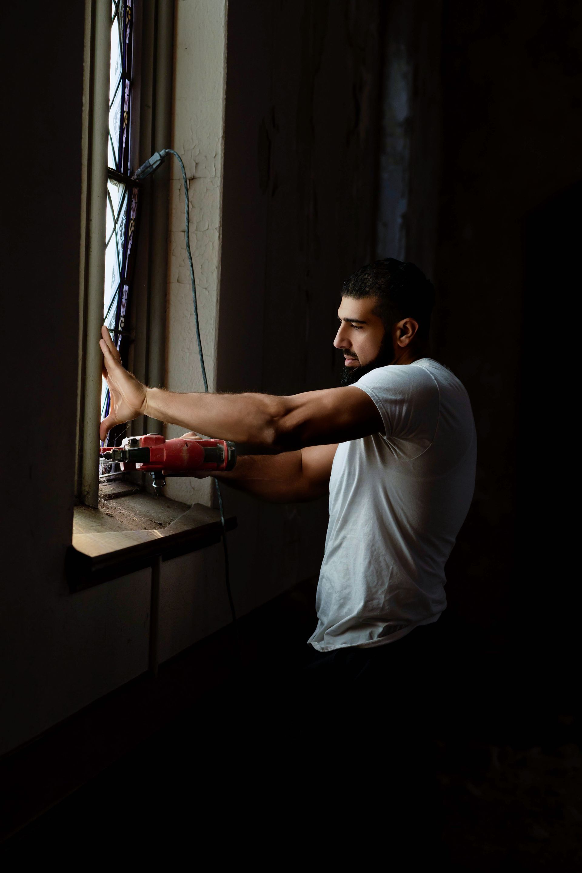 Man in white t-shirt looks out a window. Dark room, sunlight, arms outstretched.