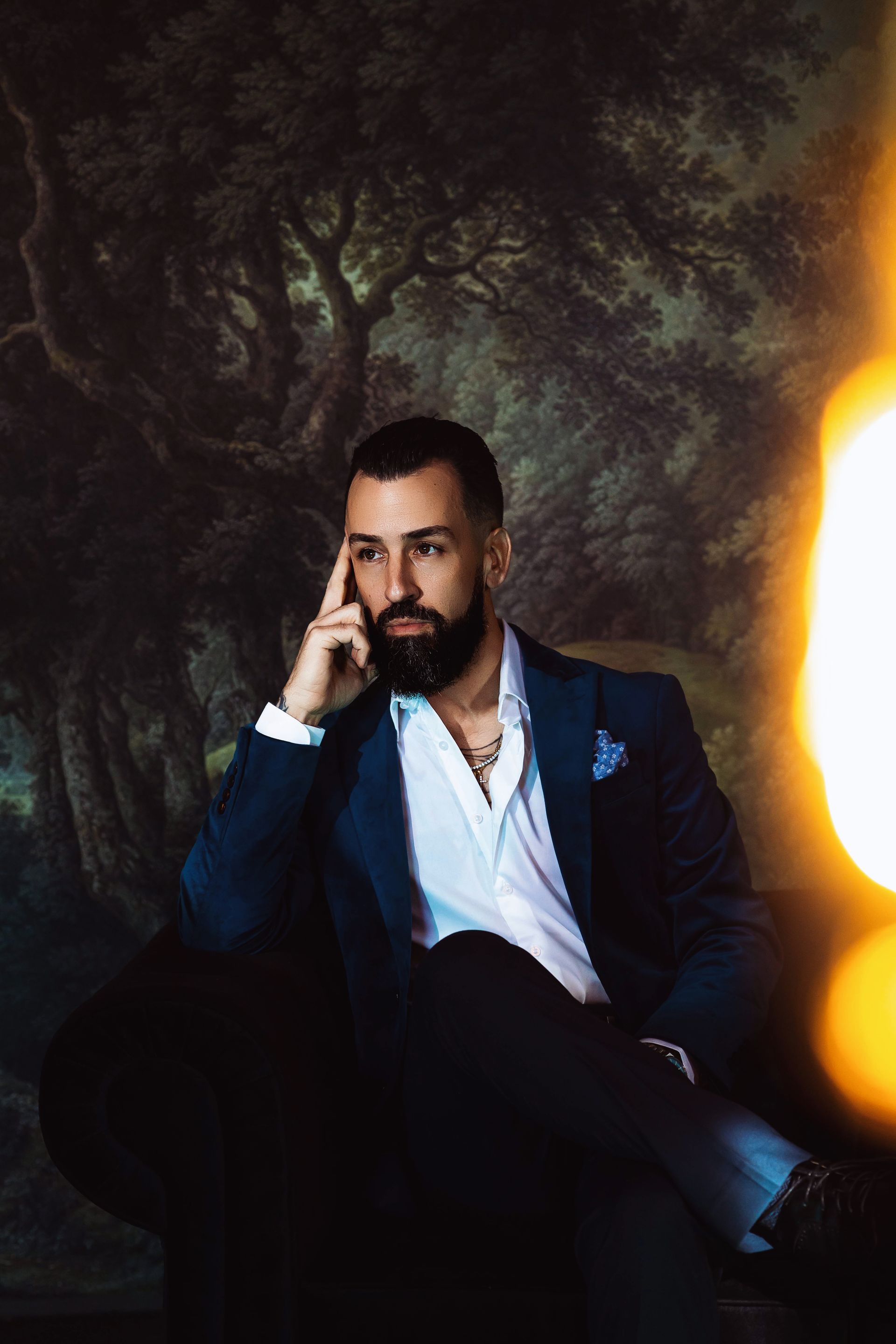 Man in navy suit, white shirt, and black pants sits thoughtfully against a dark, ornate background.