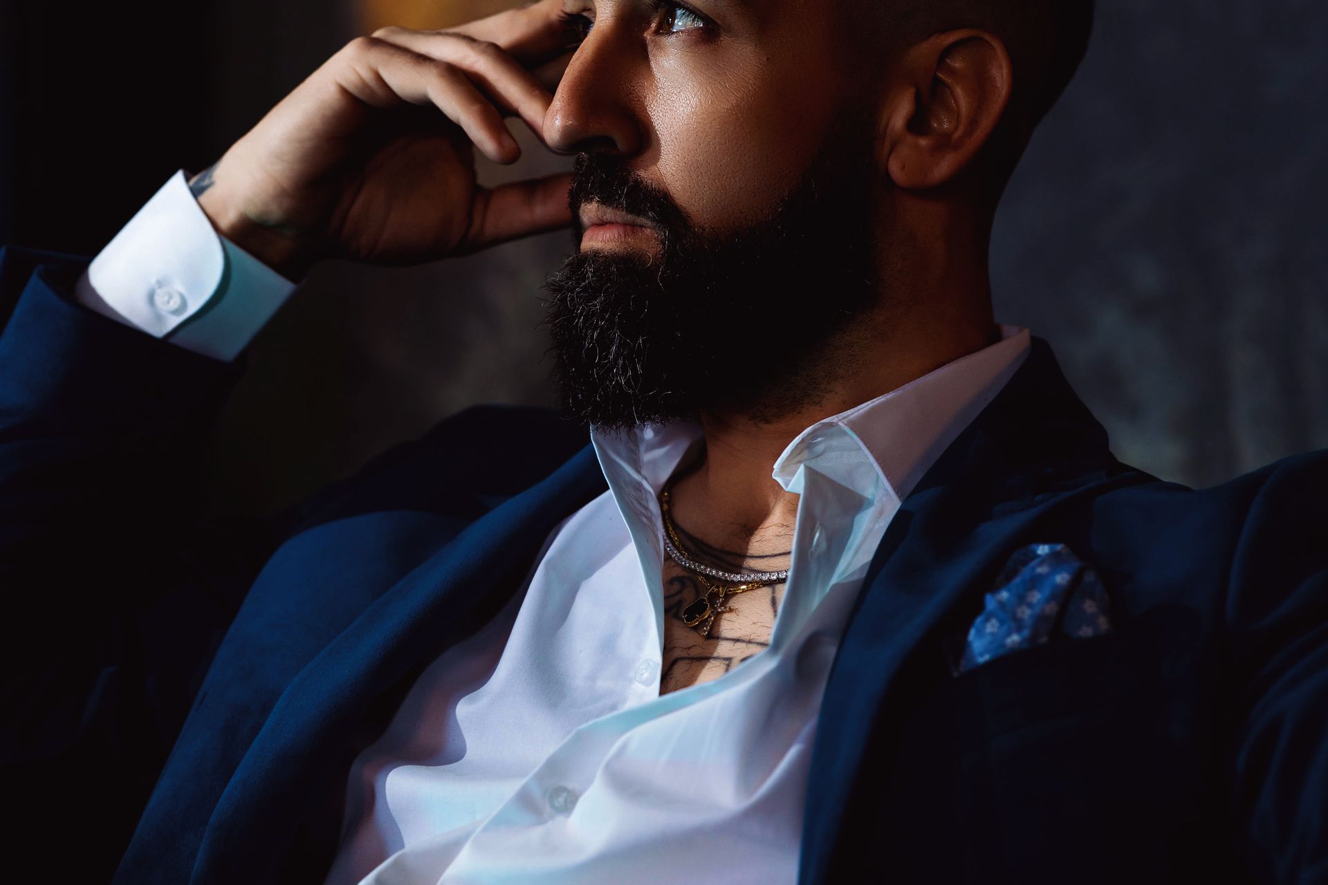 Man in a blue suit, leaning with hand on head, thoughtful expression, dark beard, white shirt.