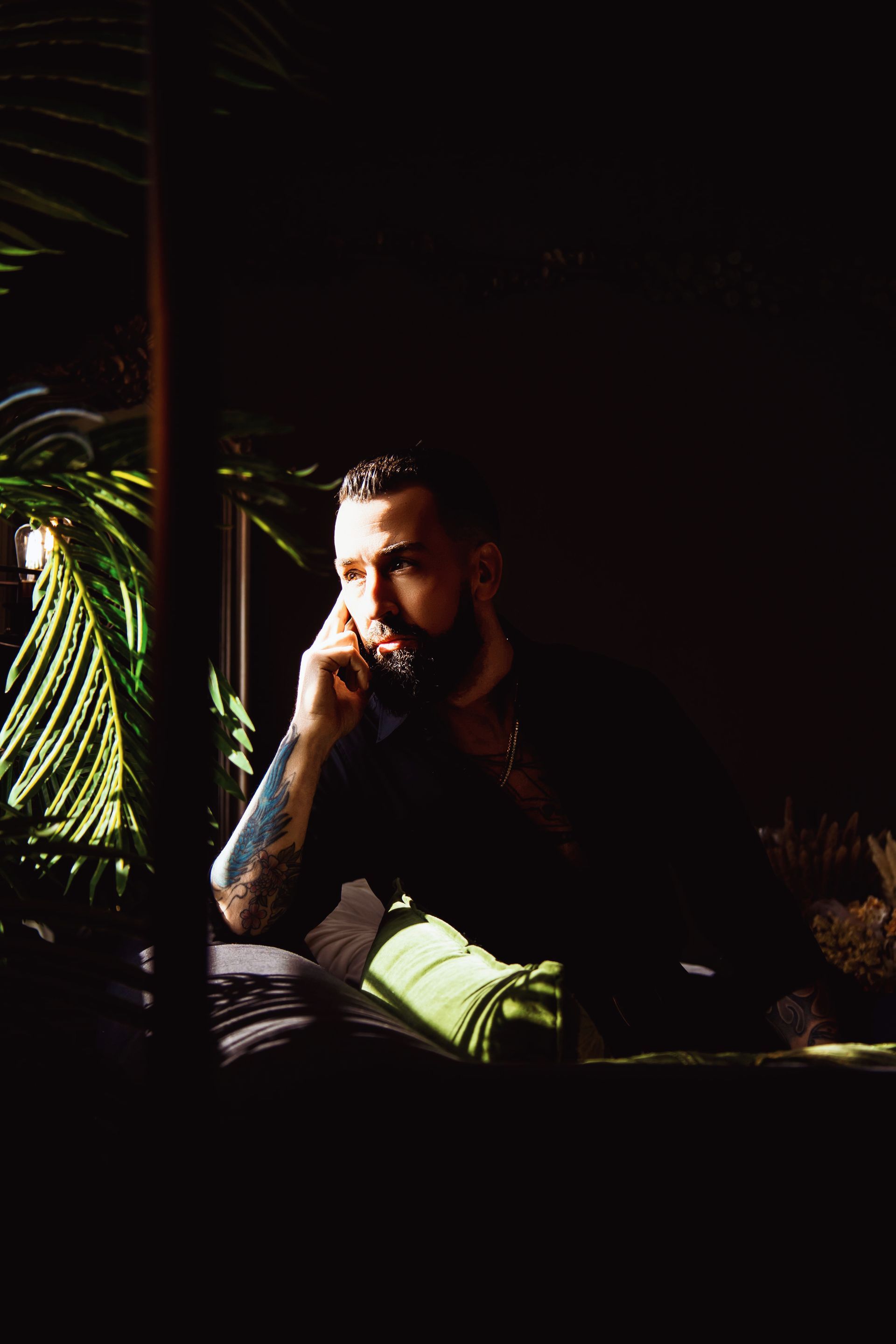 Man with beard resting chin on hand, lit by sunlight, near a green plant.