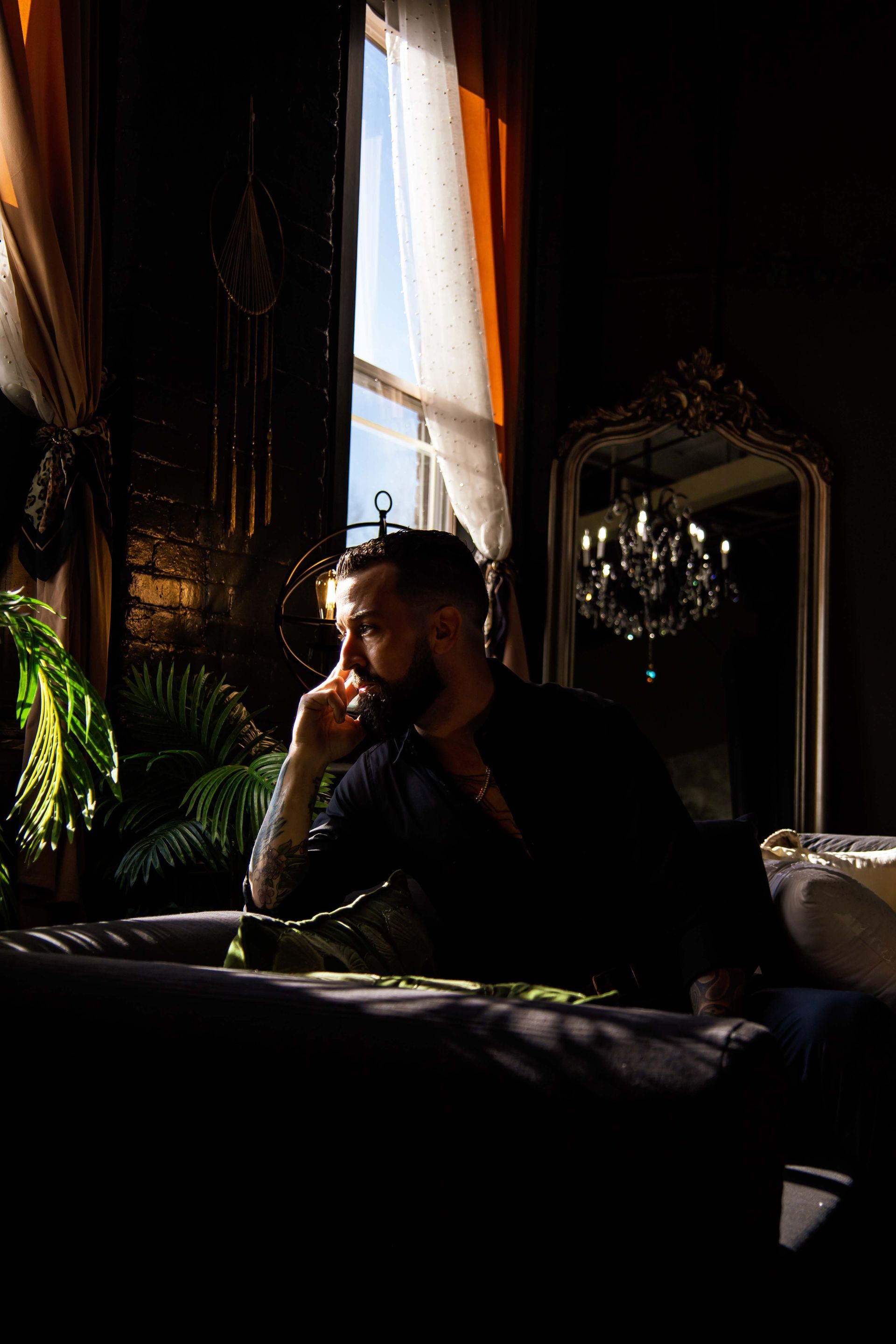 Man in dark room, resting chin on hand, looking out window. Sunlight illuminates him.