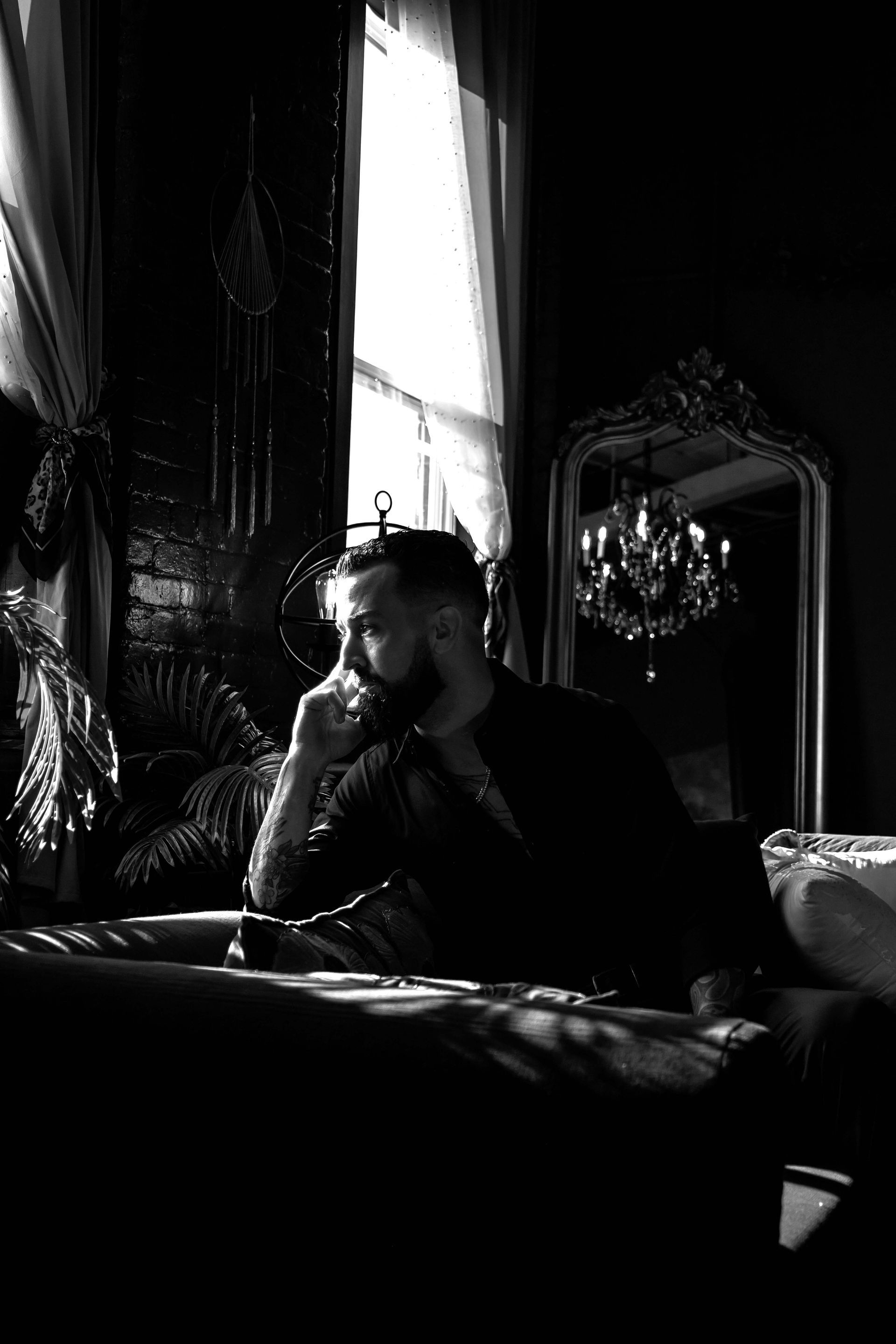 Man sits thoughtfully near window, dark room. Light streams in. Ornate mirror, chandelier visible.