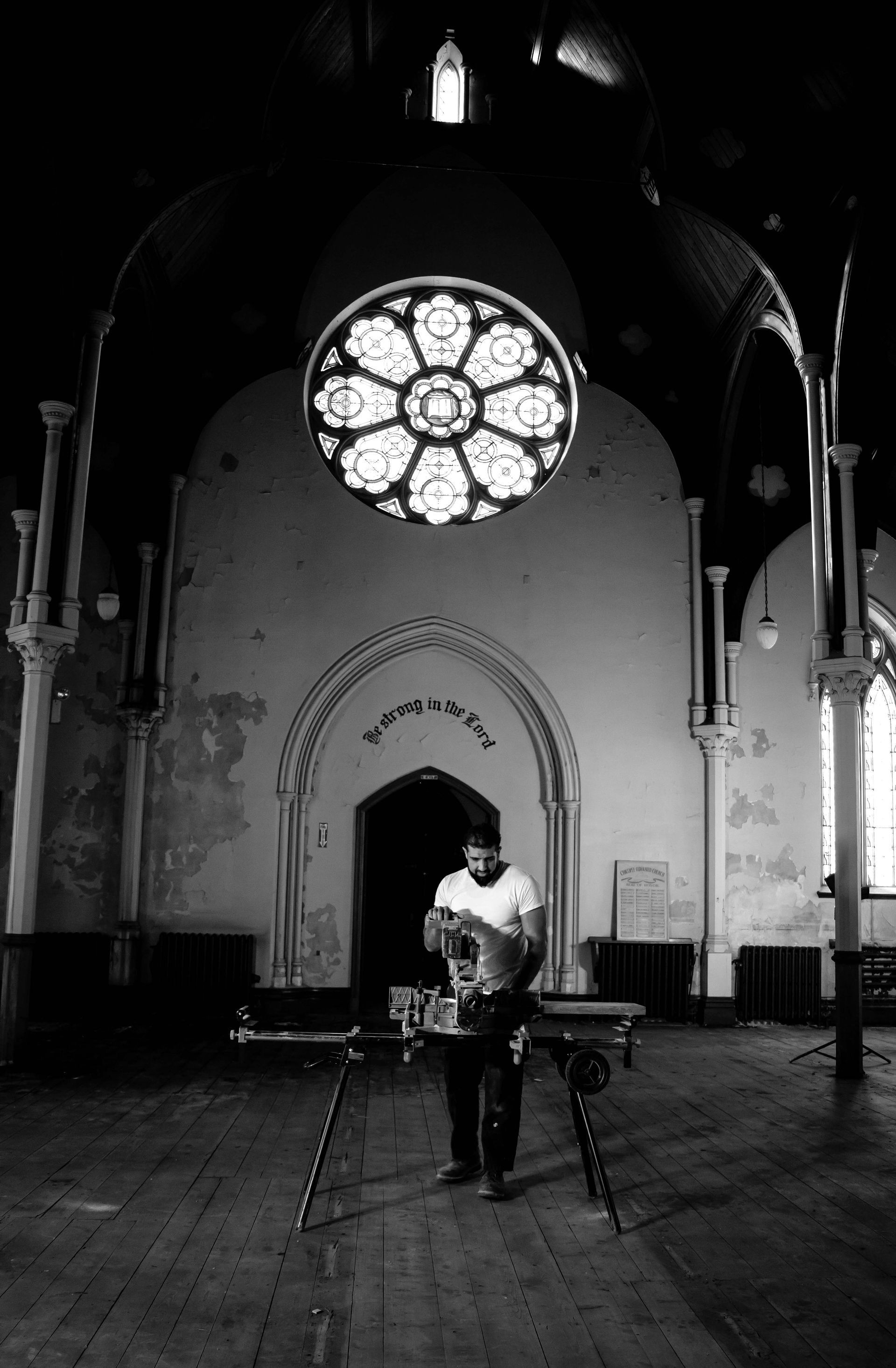 Man working at a table in a dimly lit church with a large stained-glass window above him.
