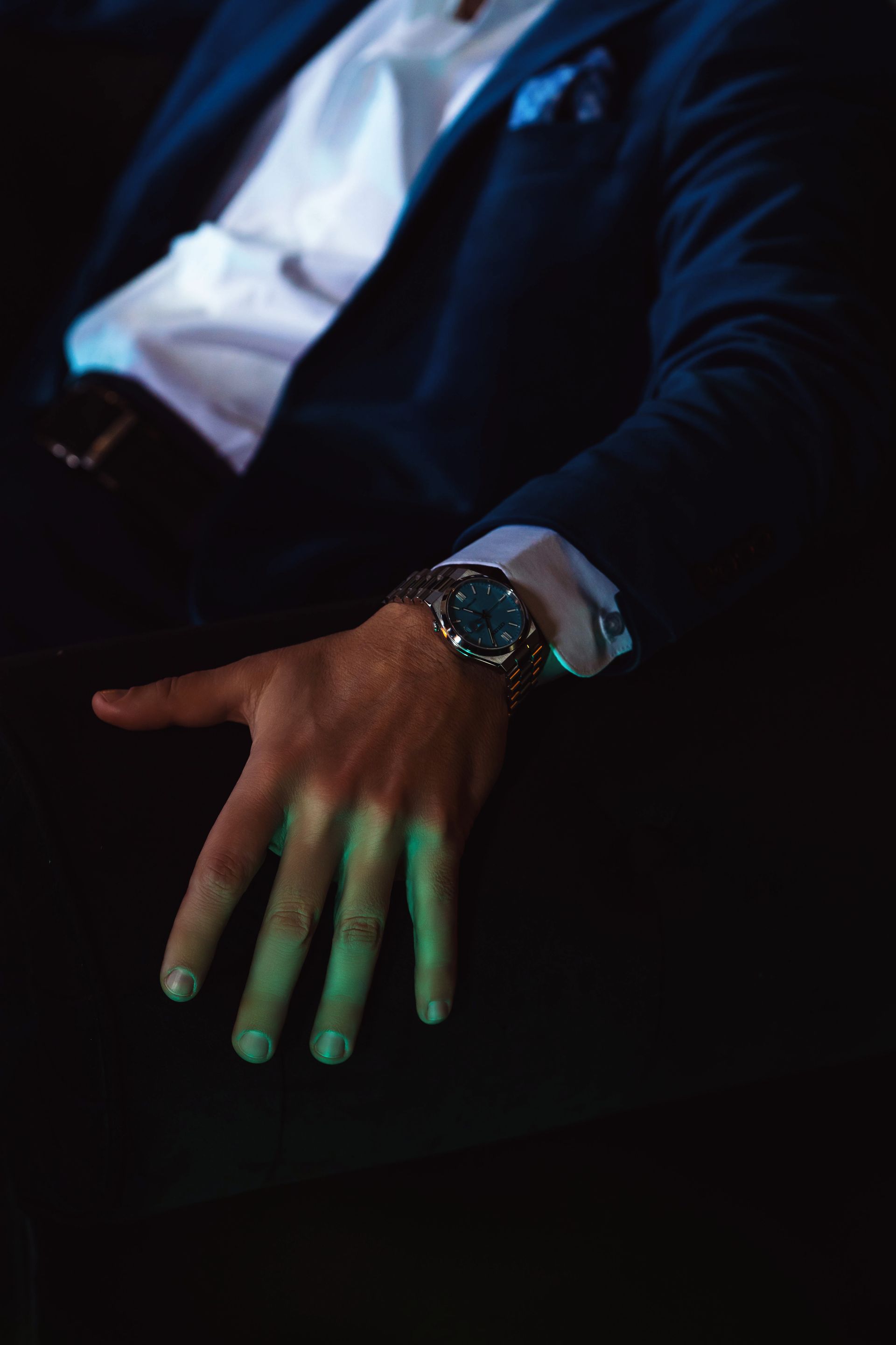 Man's hand with gold watch, resting on dark fabric; teal and blue lighting.