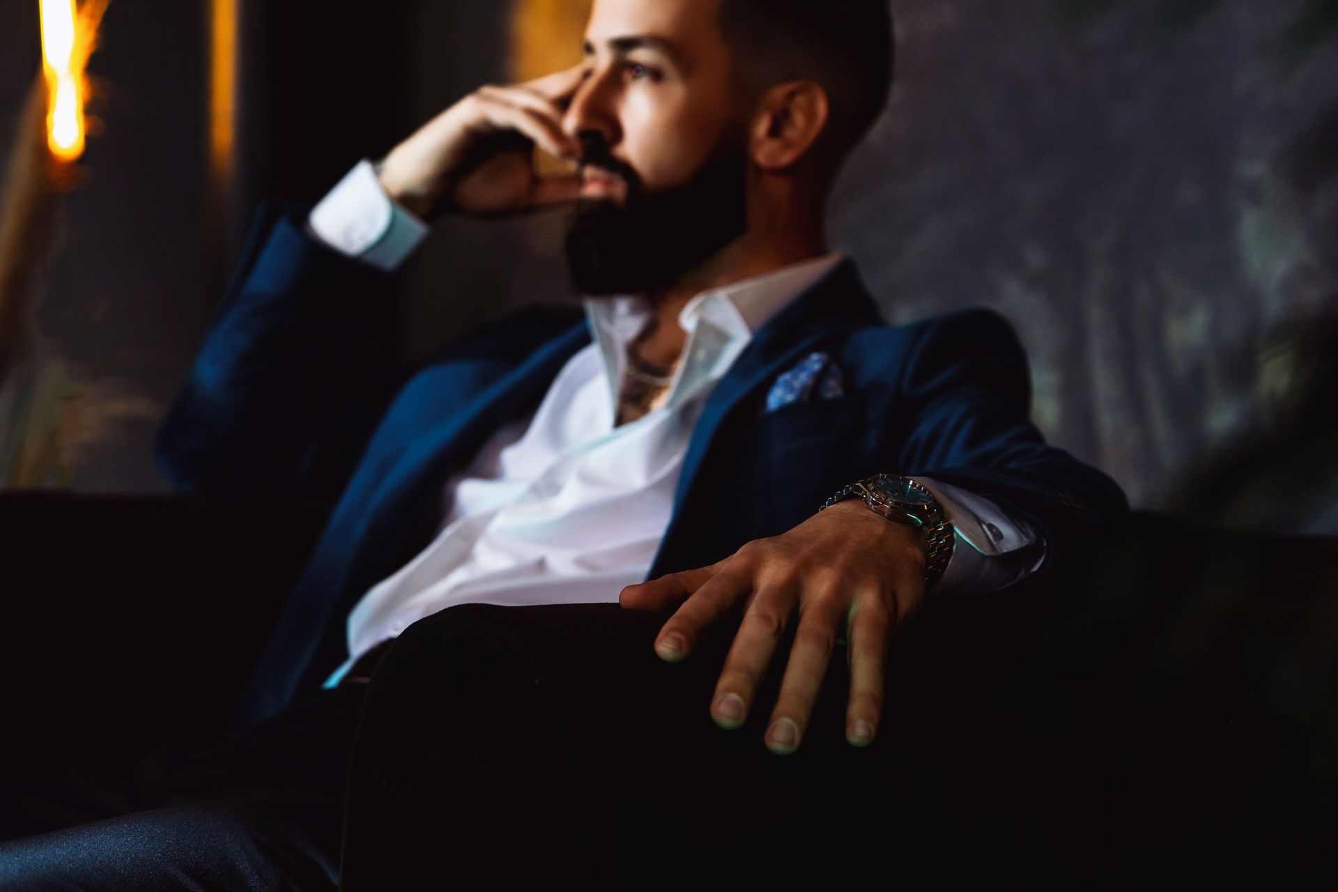 Man in a suit sits, hand on face, thoughtful expression. Dark setting.