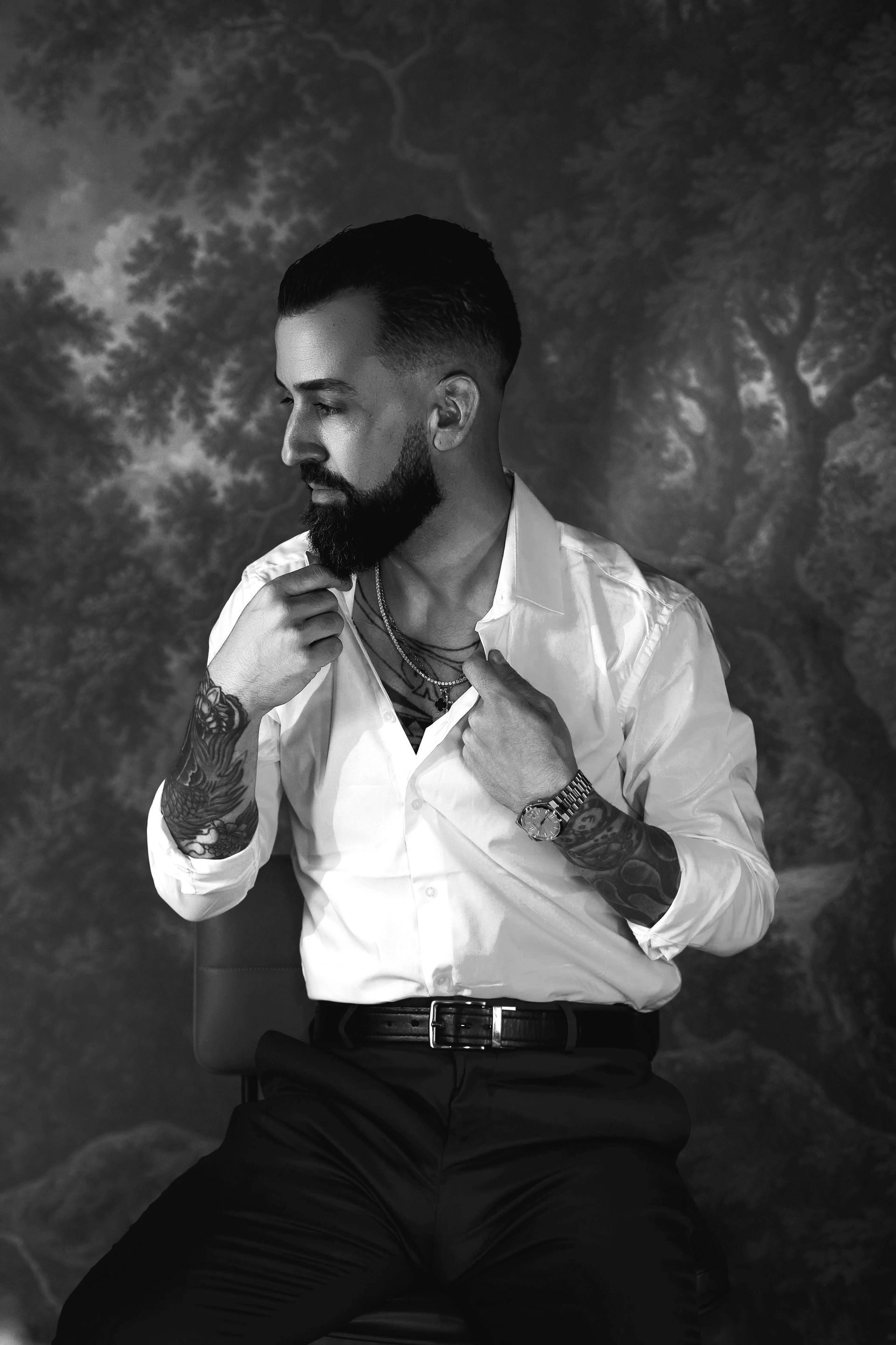 Man with tattoos adjusts his shirt, posing against a landscape backdrop.