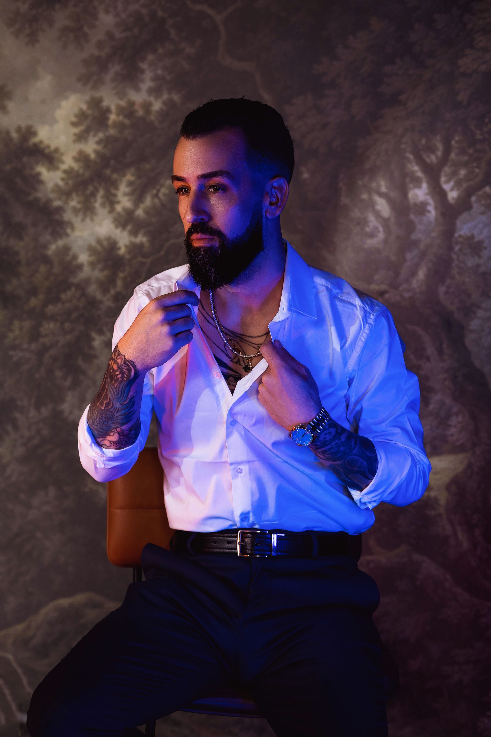 Man in white shirt, seated, adjusting shirt, tattoos visible. Blue lighting, floral backdrop.