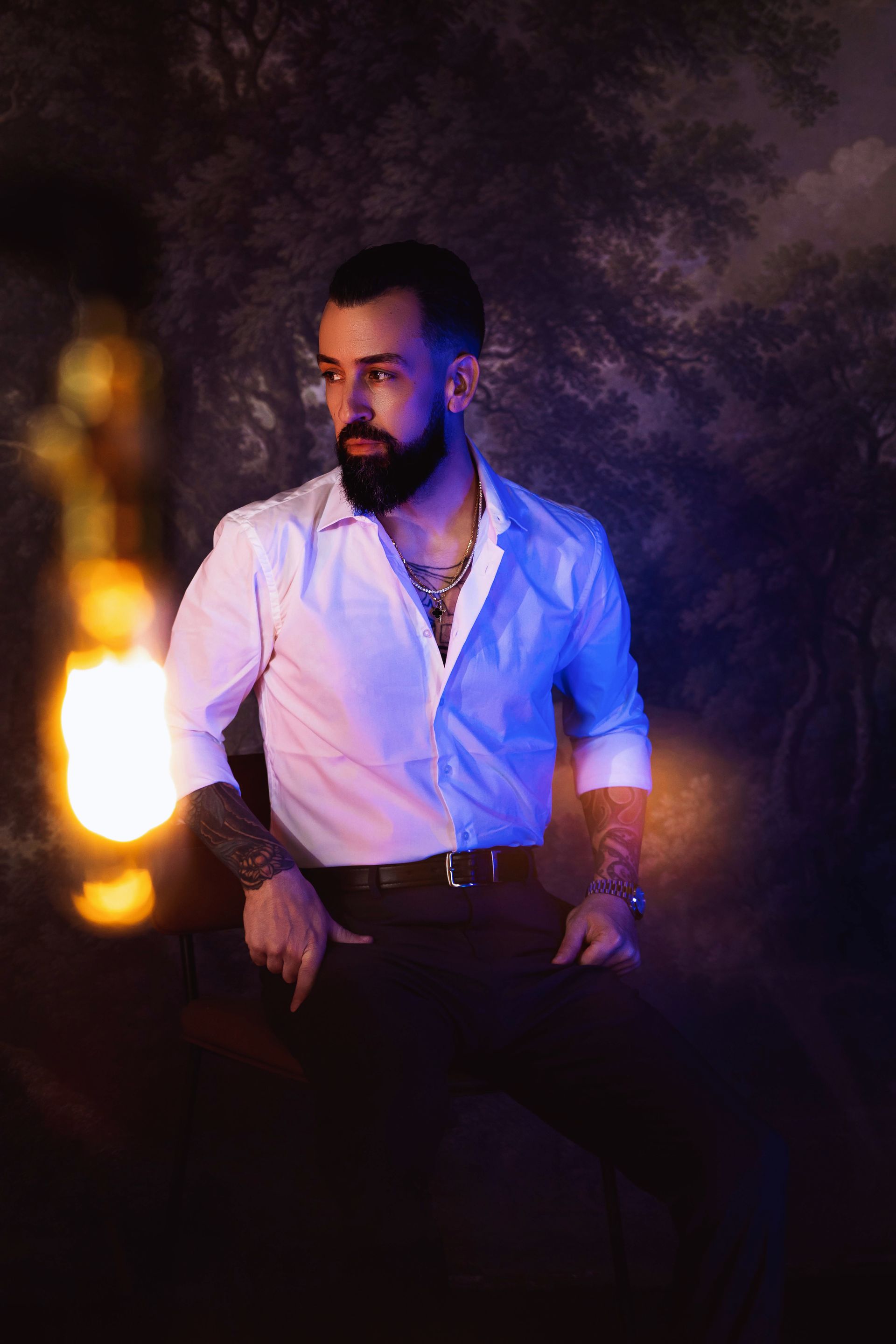 Man with beard and tattoos in a white shirt, holding hand in pocket, stands lit by warm and cool light.