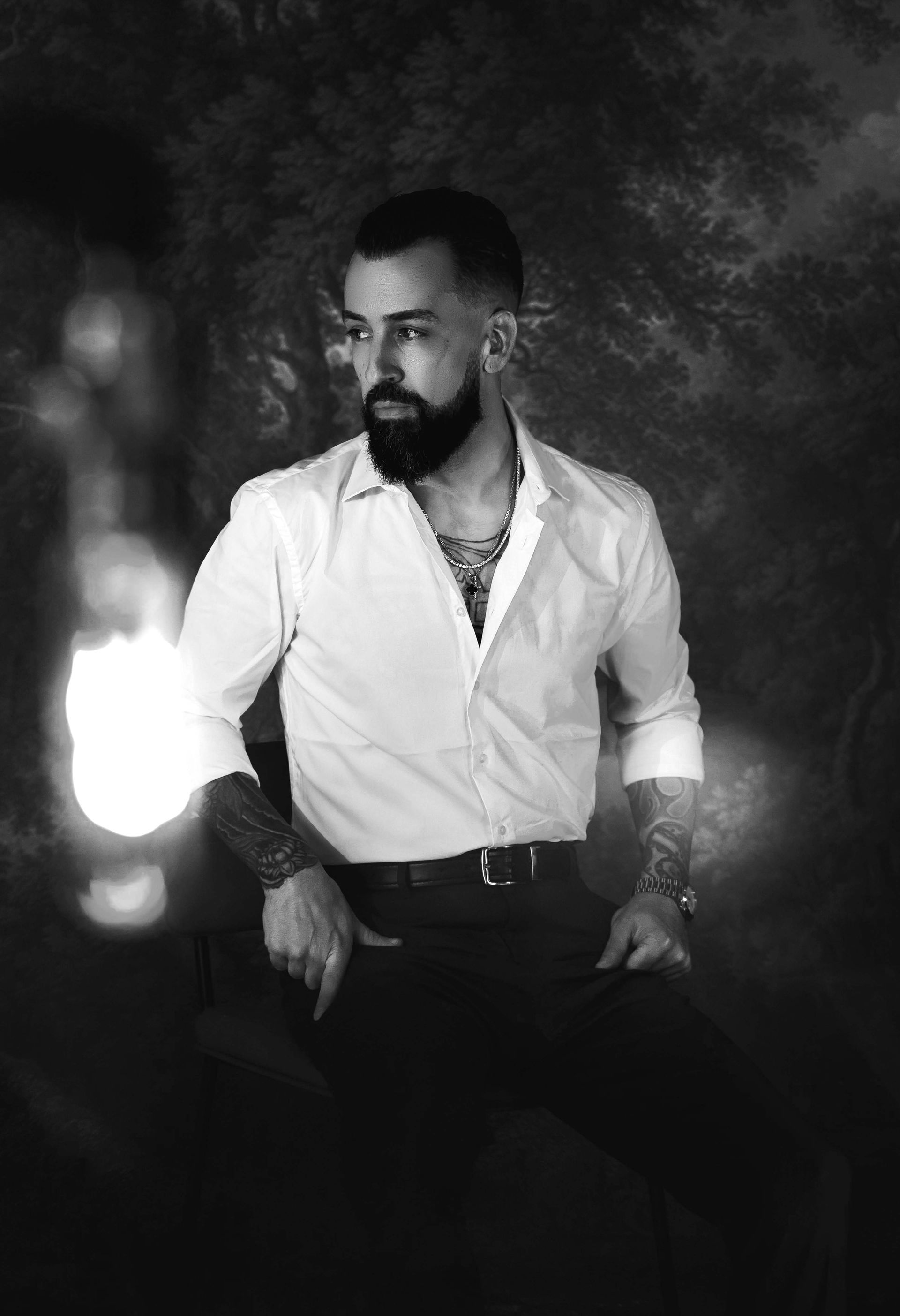 Man with beard, tattoos, white shirt, black pants, looking away, dark backdrop.